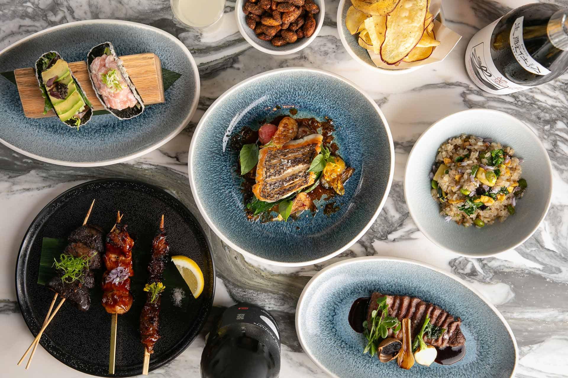 A variety of gourmet dishes are artfully arranged on a marble table. There are skewers with meats, bowls of diced vegetables and grains, grilled fish, chips, nuts, and a bottle of wine. The presentation is elegant, featuring ceramic plates.