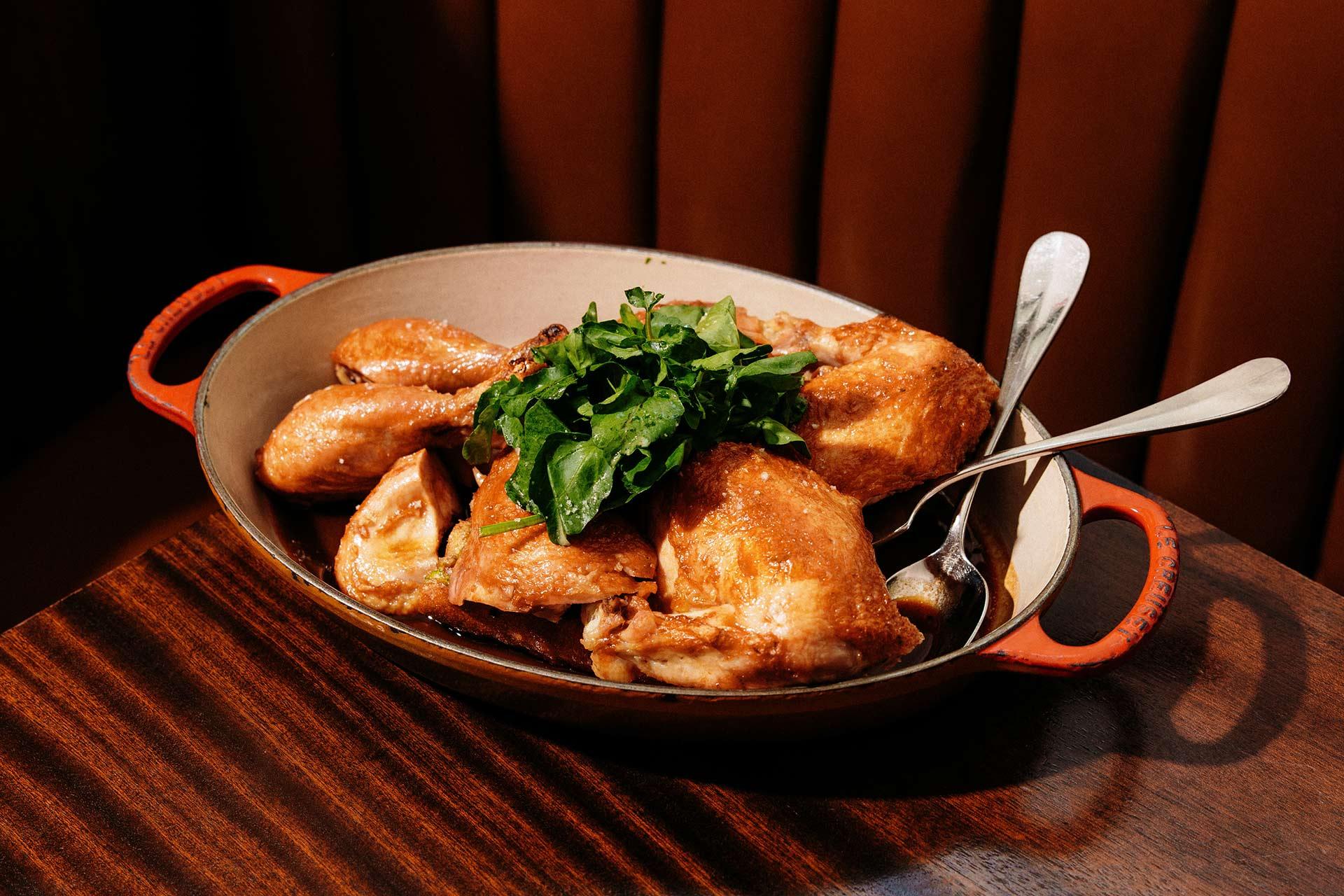 A baked or roasted chicken cut into pieces is served in an oval dish, garnished with fresh greens, with serving utensils placed inside. The dish sits on a wooden table.