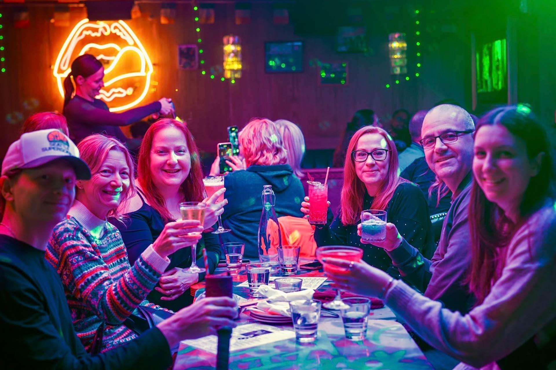 A group of six people sit around a table in a colorful bar, smiling and raising drinks. Neon lips sign glows on the wall behind them. The atmosphere is lively and festive, with bright lights and decorations.