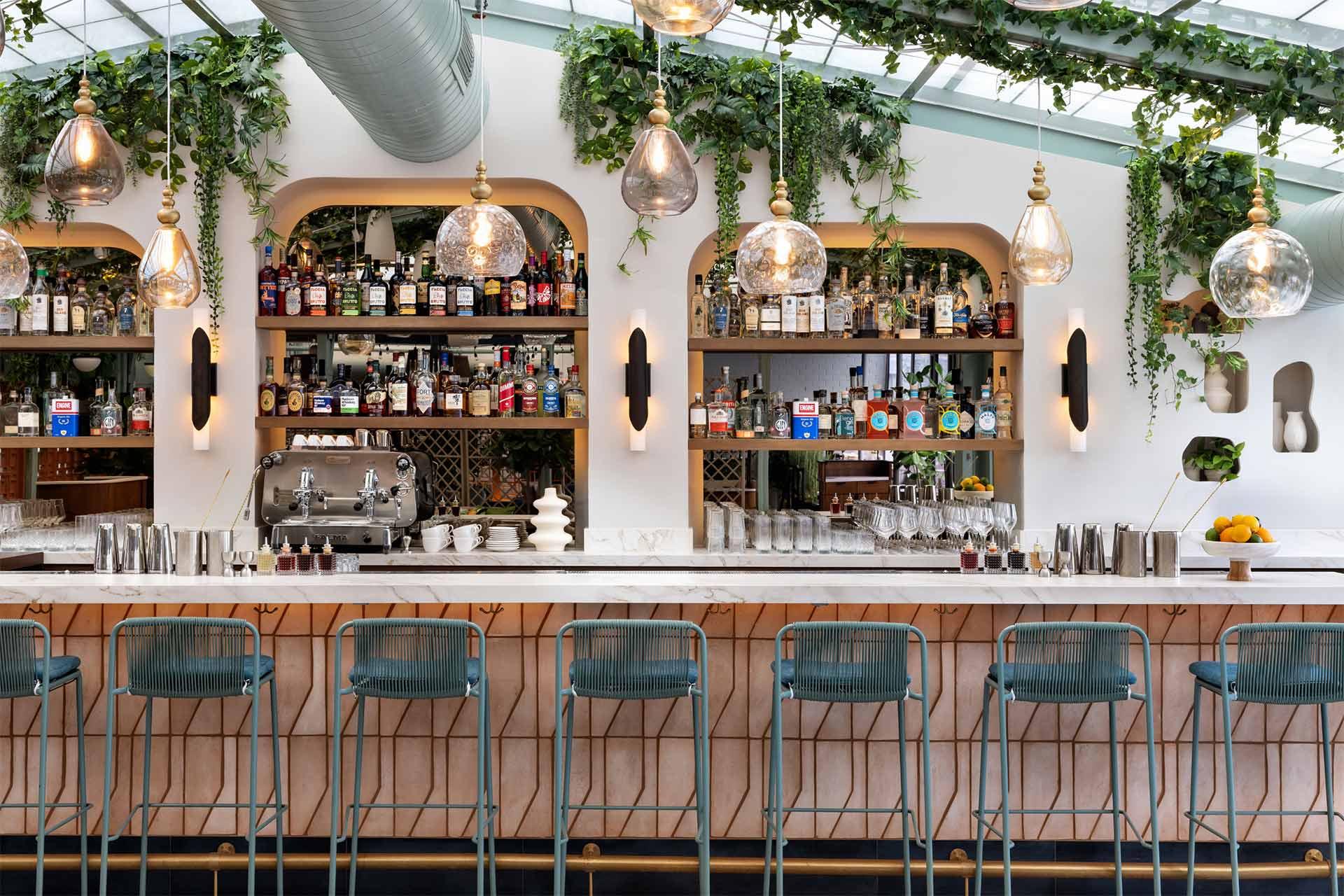 A modern bar with a marble countertop, teal metal bar stools, hanging glass pendant lights, and shelves stocked with bottles. Green plants and vines decorate the upper shelves and white walls.