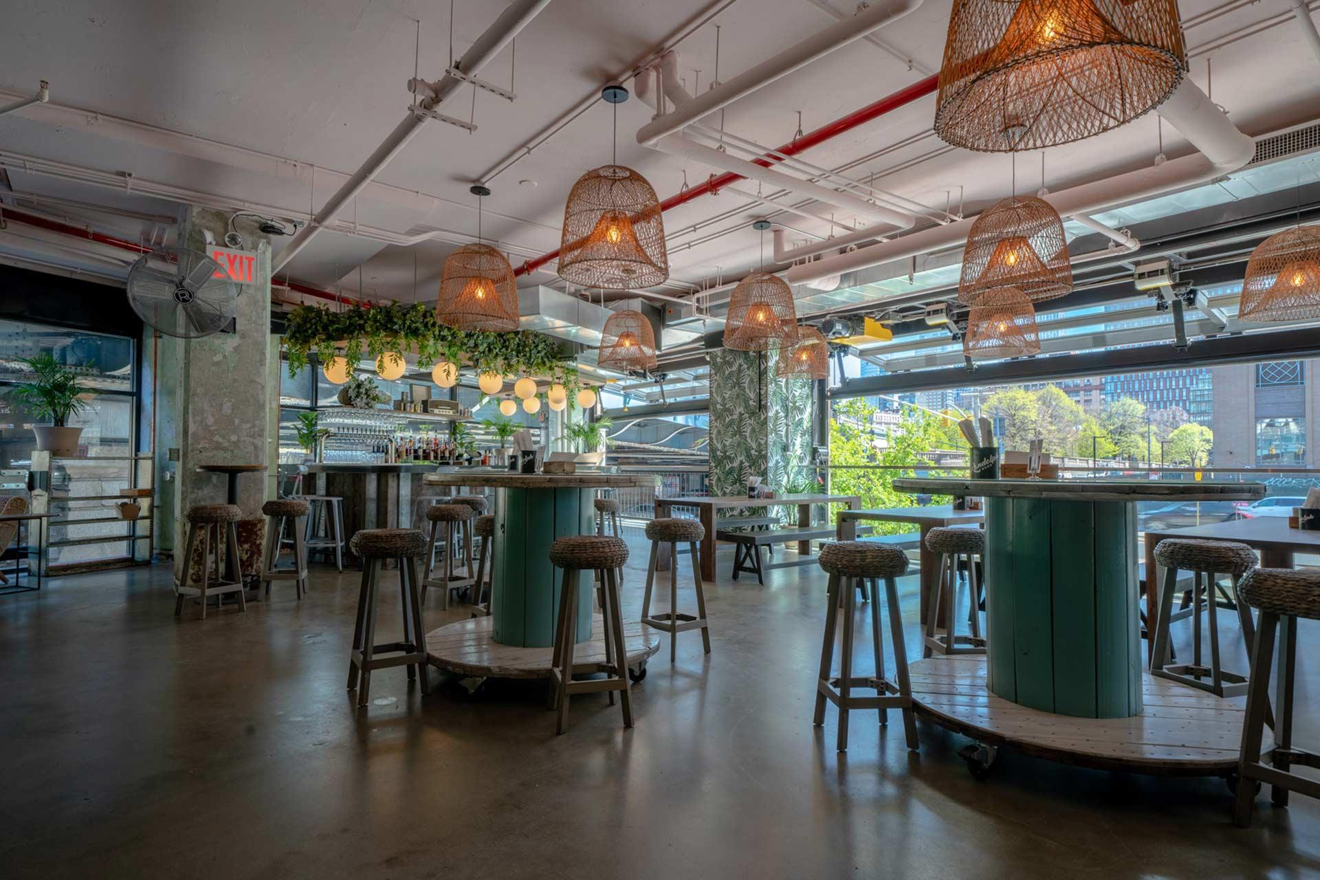 Spacious, modern bar with high tables and stools, wicker pendant lights, hanging plants, large windows with city views, and natural light filling the room.