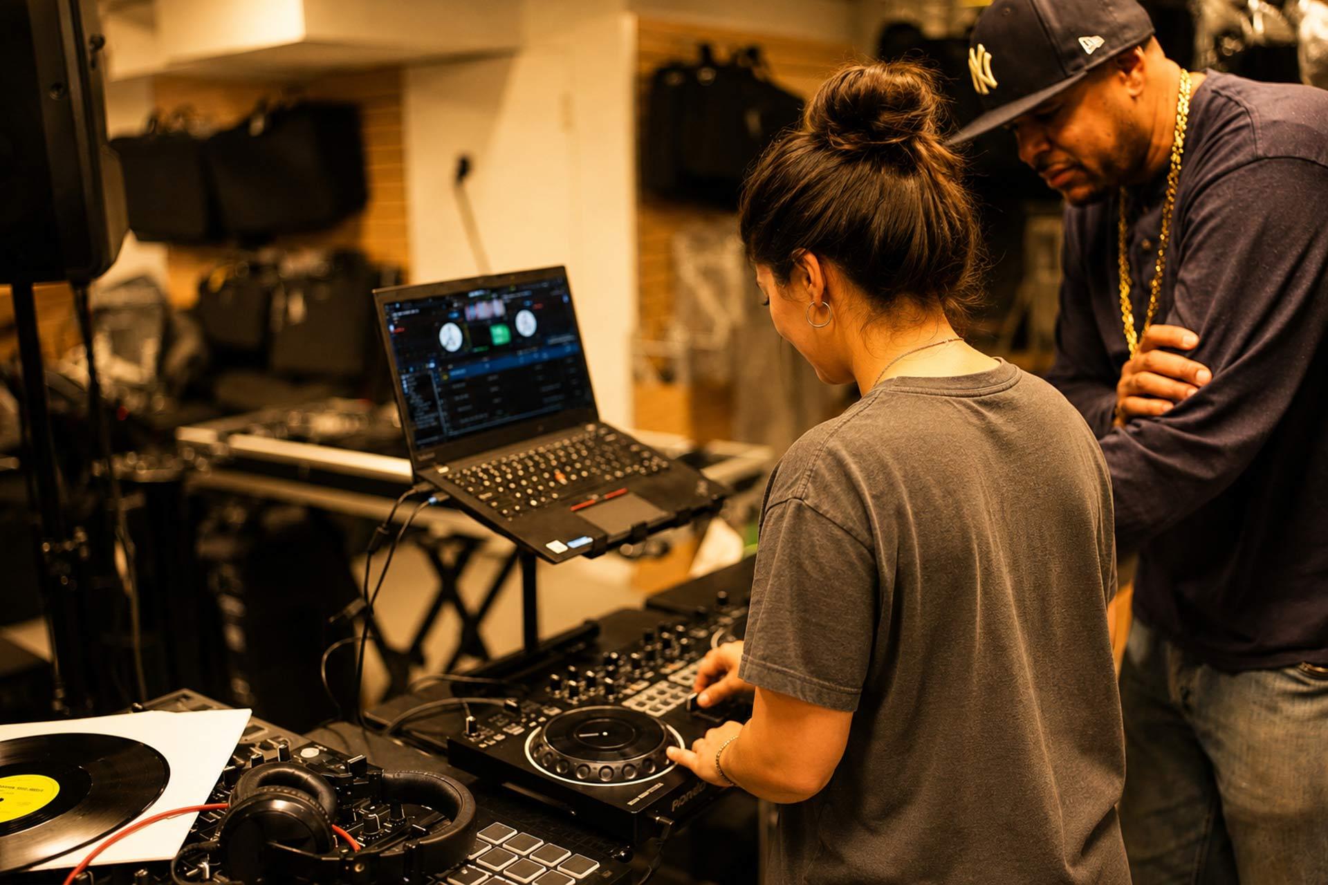 A woman operates DJ equipment and a laptop while a man wearing a cap and gold chain looks on in a music studio filled with audio gear and instruments.