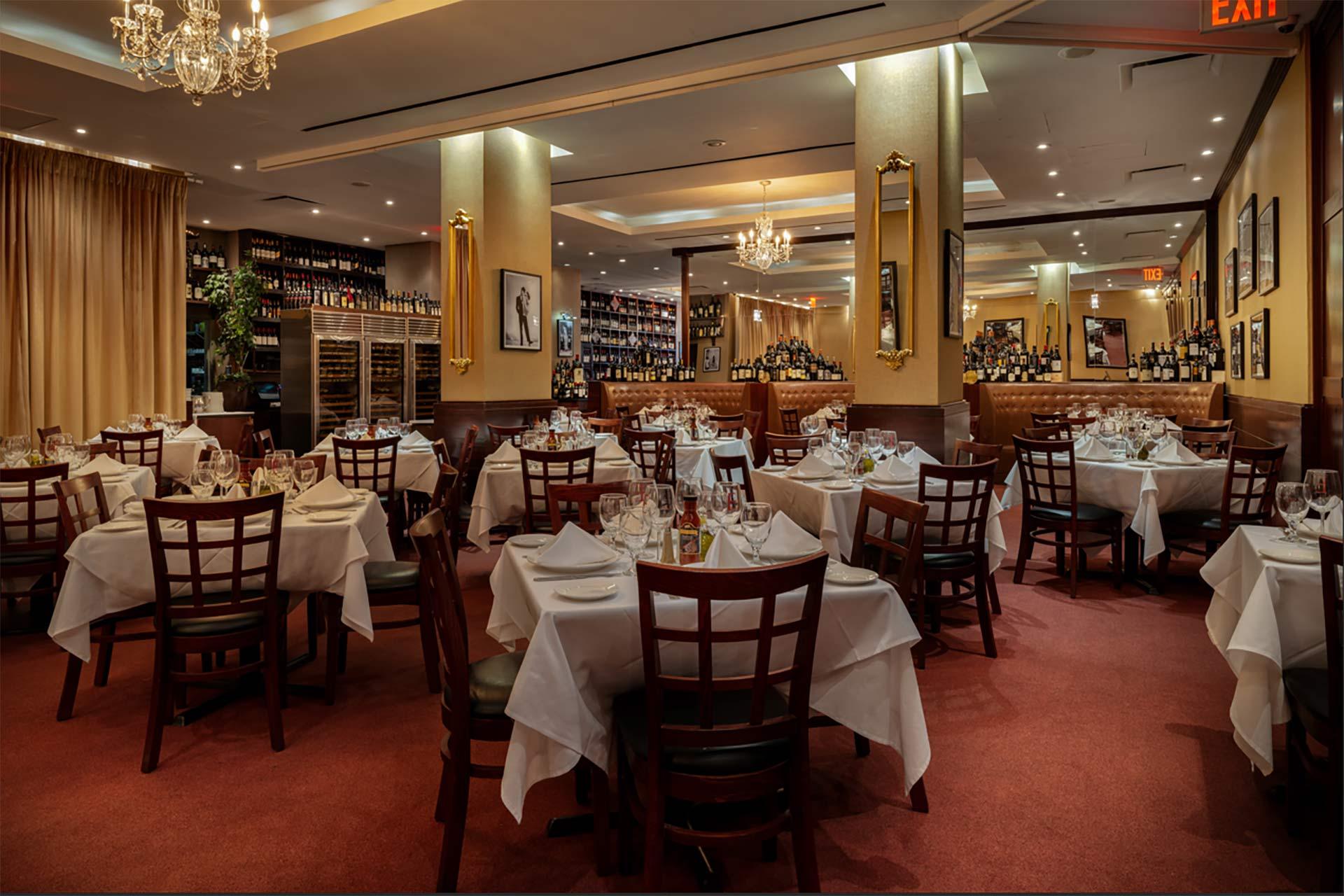 A warmly lit restaurant dining room with round tables covered in white tablecloths, set with glassware and plates. Wooden chairs surround the tables, and wine bottles line shelves on the walls. Chandeliers hang from the ceiling.