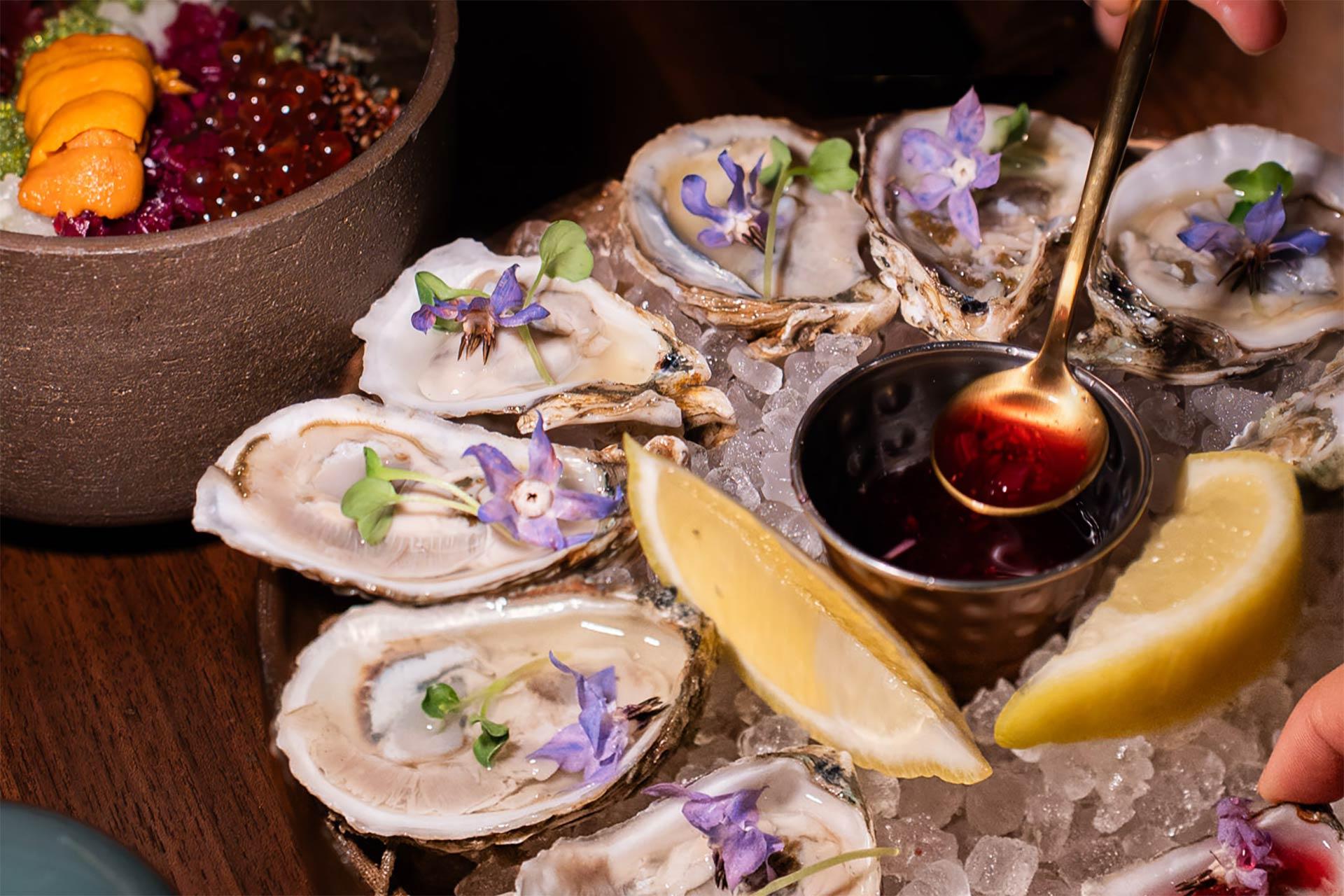 A platter of fresh oysters on ice garnished with edible flowers, lemon wedges, and microgreens. A hand holds a spoon over a small cup of sauce, ready to pour it onto the oysters. A bowl of uni and roe is in the background.