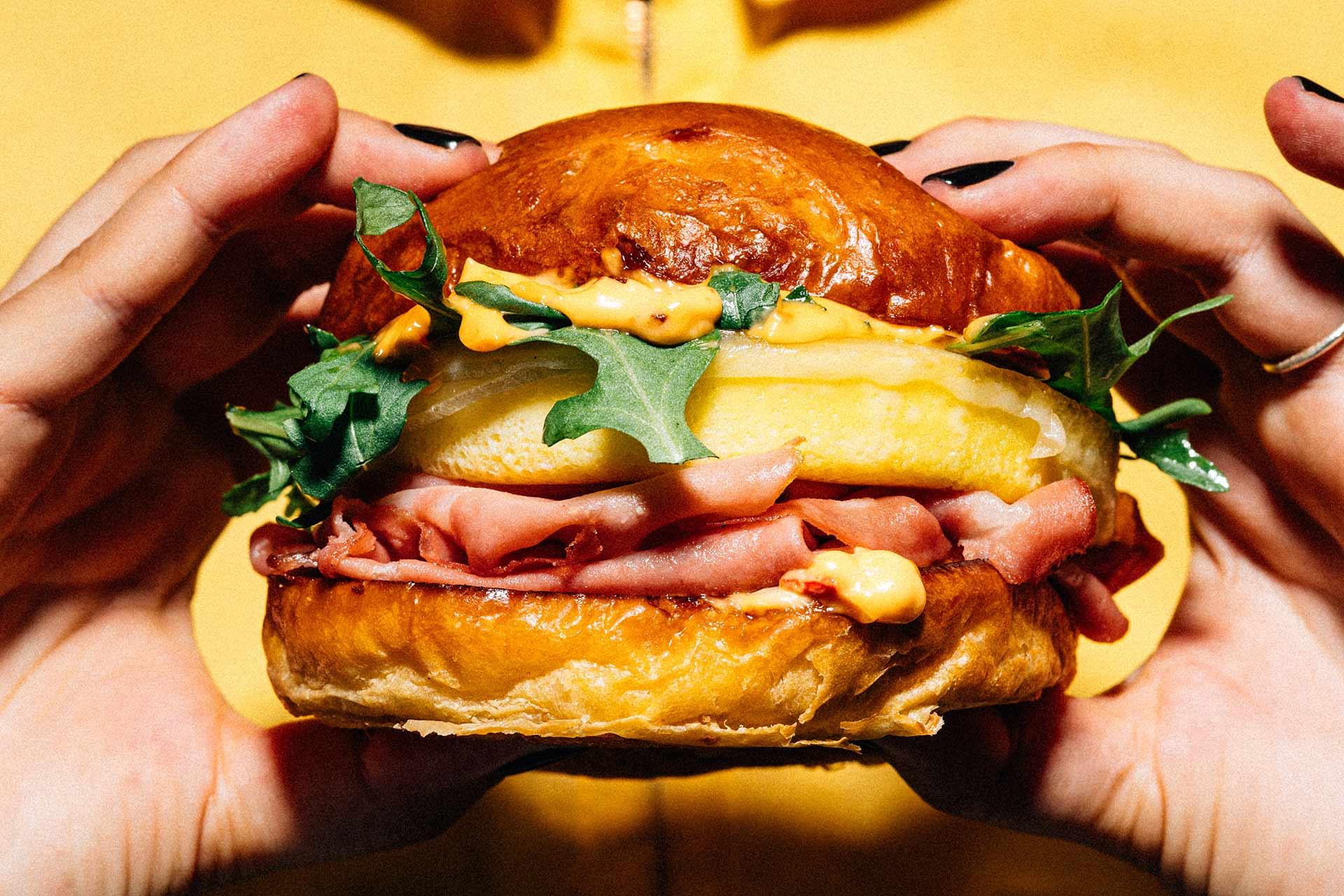 A person holds a sandwich with a shiny brioche bun, layers of ham, melted cheese, arugula, pineapple, and a drizzle of orange sauce, against a yellow background.