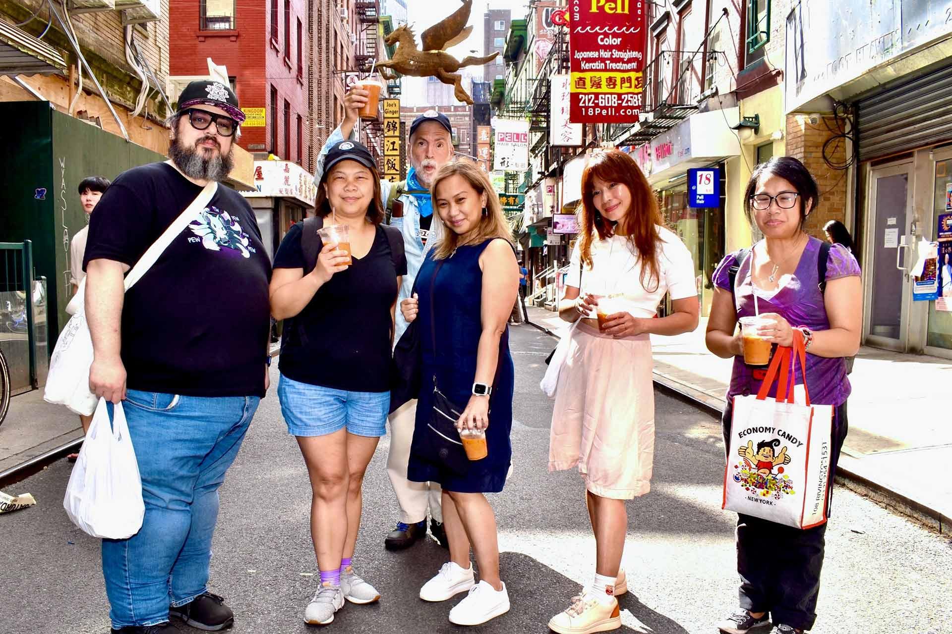 Six people stand together on a city street holding drinks, posing for a group photo. They are casually dressed and smiling, with shops, signs, and buildings visible in the background.