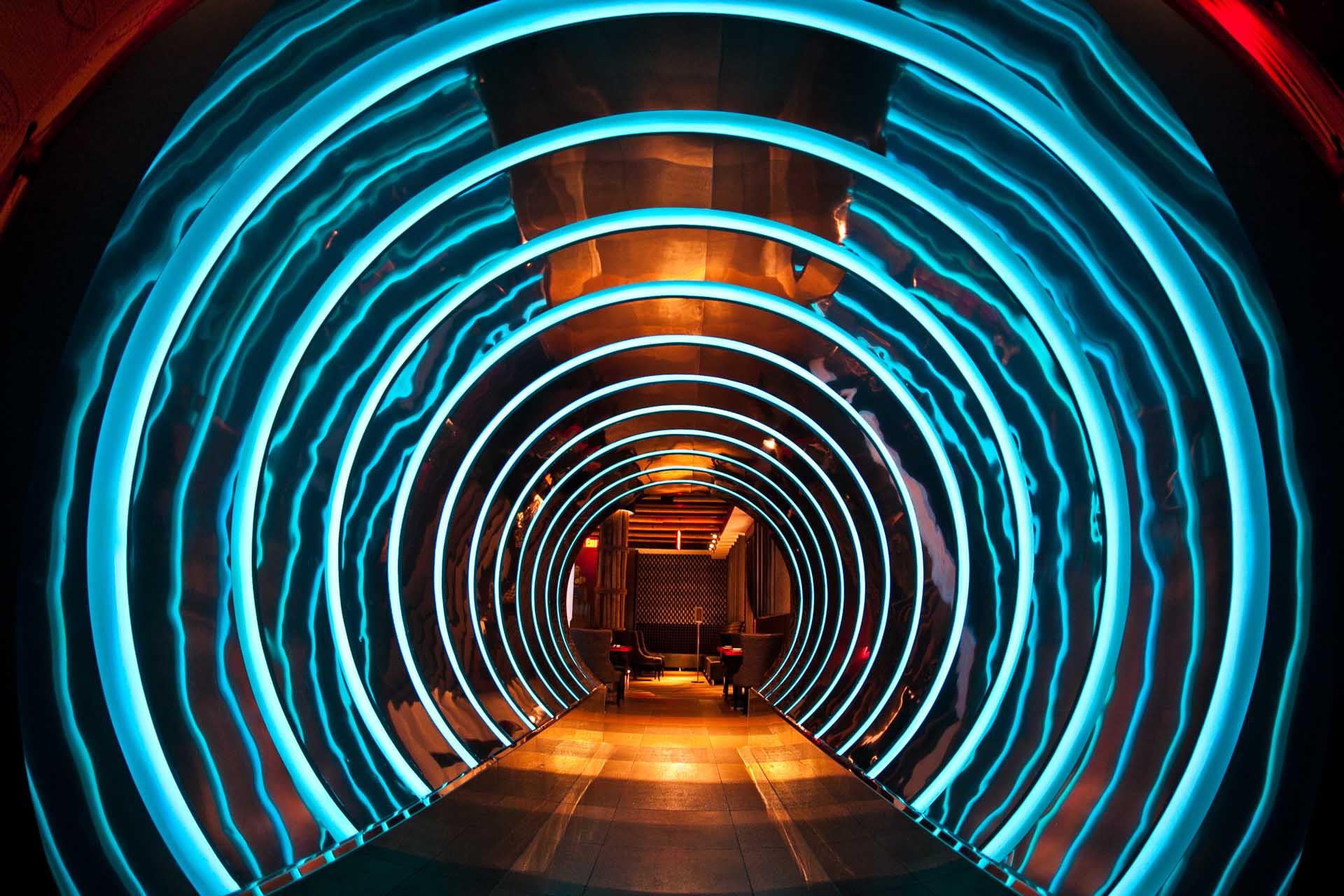 A futuristic hallway features concentric blue neon rings illuminating the tunnel, leading to a cozy seating area with chairs and warm lighting at the far end. The walls and floor have a metallic, modern appearance.