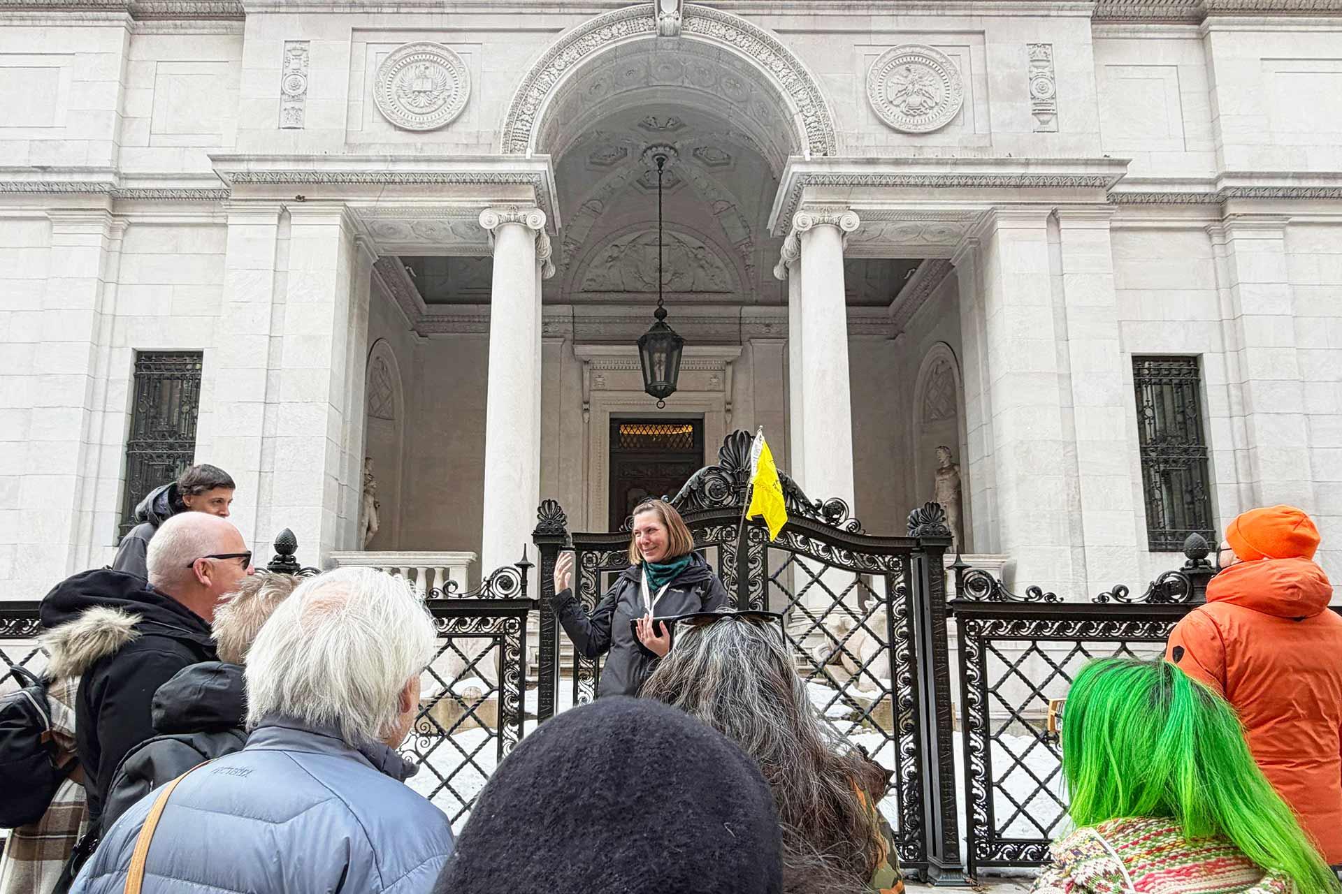 A tour guide speaks to a group of people gathered outside a grand, historic building with tall columns and ornate architecture. One person in the crowd has bright green hair, and another holds a yellow flag.