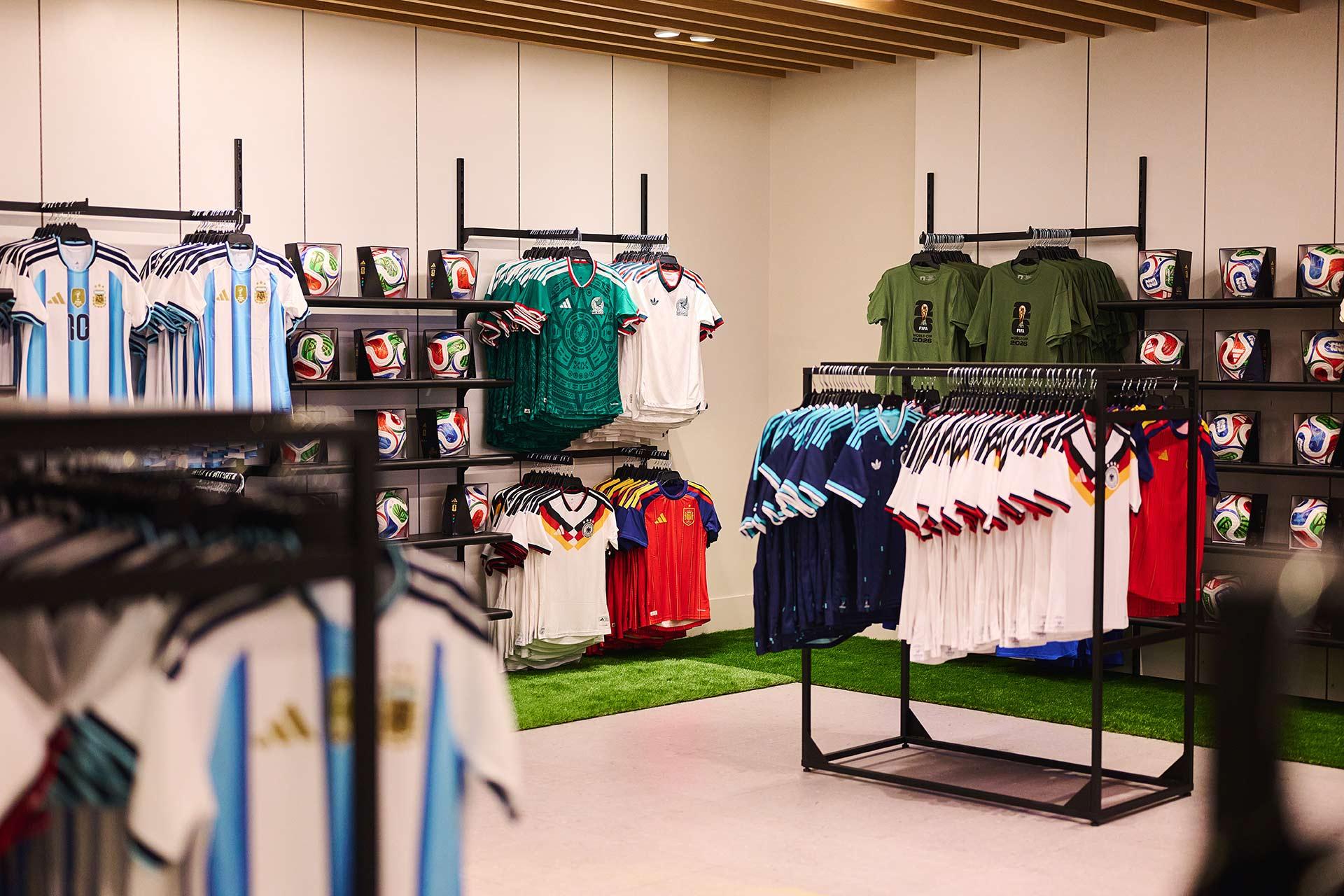 A sports store displays various national soccer team jerseys, including Argentina, arranged on racks. Shelves in the background hold multiple soccer balls in boxes, and the floor features patches of green artificial turf.