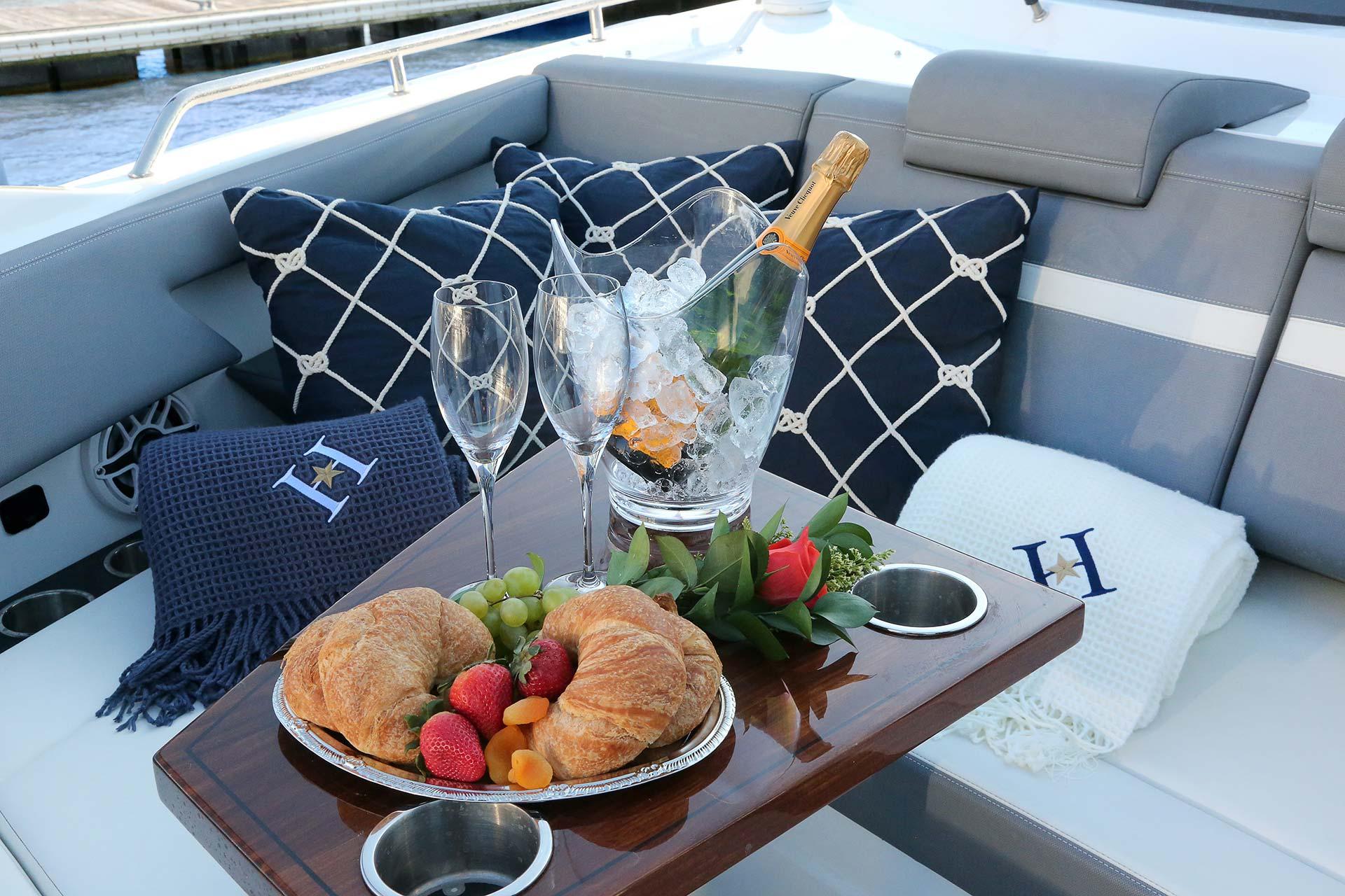 A table on a yacht displays croissants, strawberries, grapes, and apricots on a platter, with two empty champagne flutes and a bottle of champagne in an ice bucket. Nautical pillows and blankets decorate the seating area.