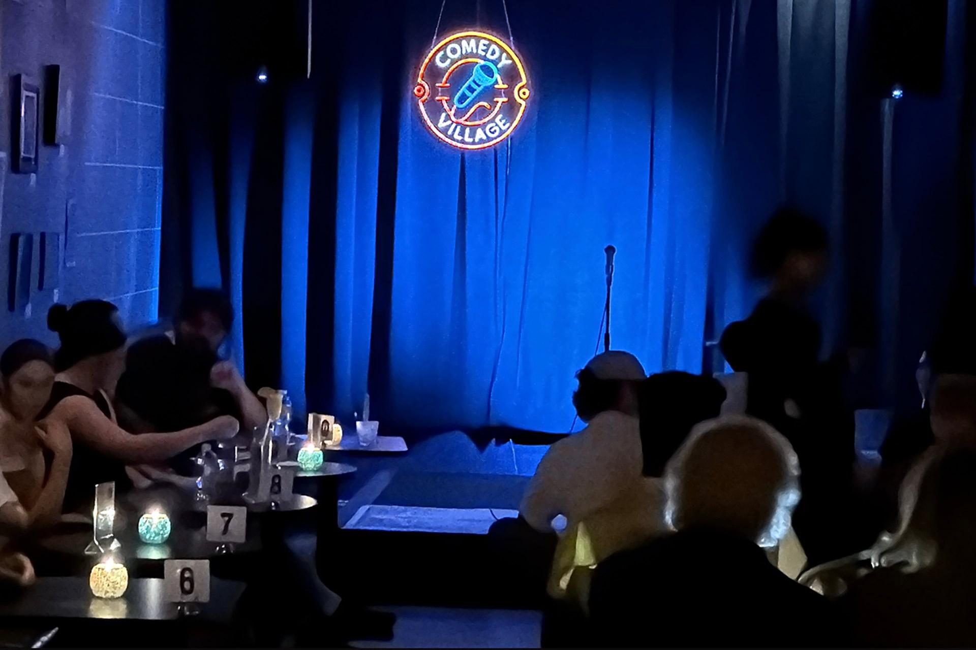 A dimly lit comedy club with a small stage, microphone stand, and a blue curtain. The neon sign above reads "Comedy Village." Audience members sit at tables, waiting for the show to begin.