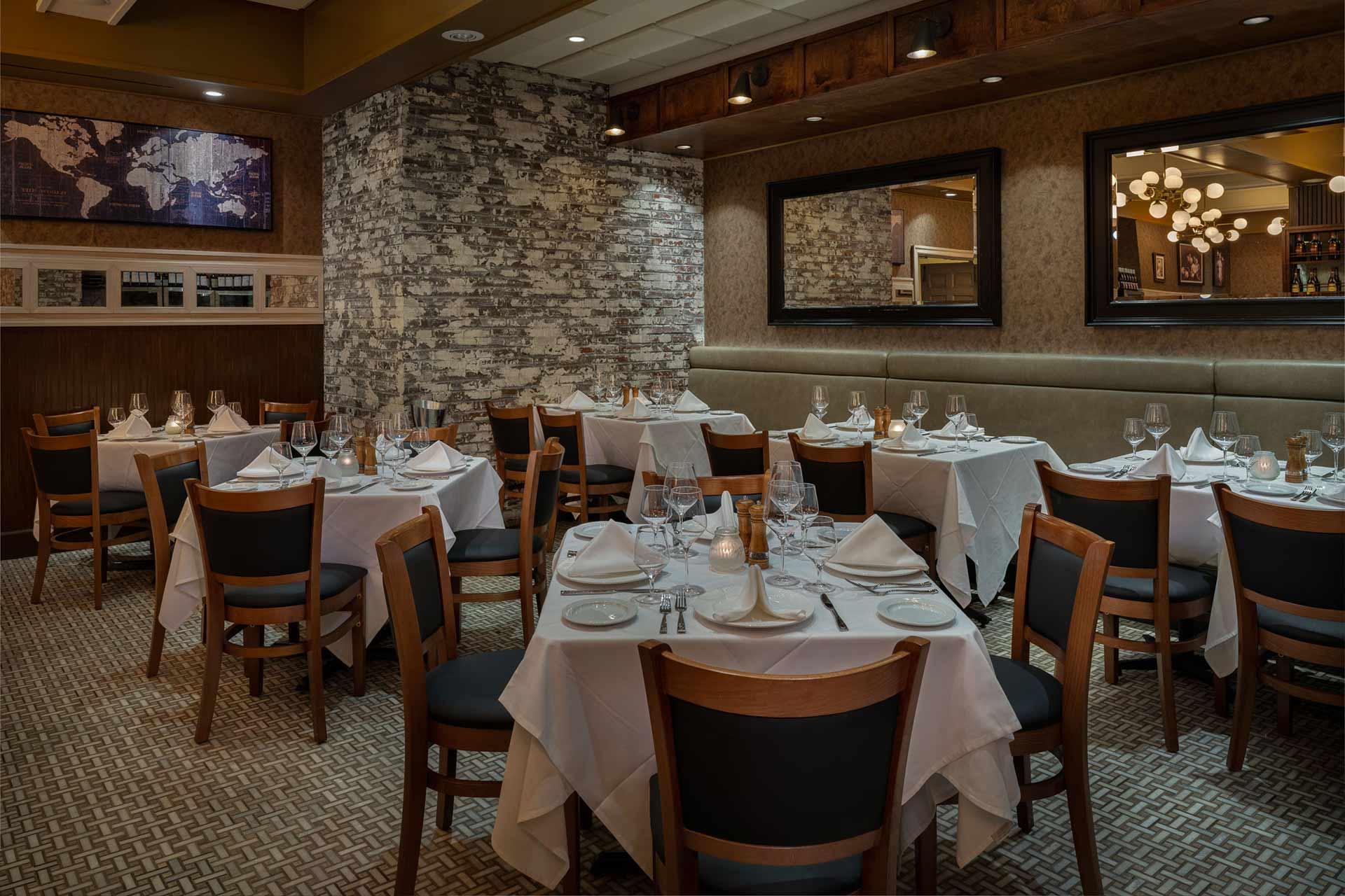 Elegant restaurant dining room with neatly set tables covered in white tablecloths, wooden chairs, exposed stone accent wall, warm lighting, mirrors, and a map on the wall, creating an inviting and upscale atmosphere.