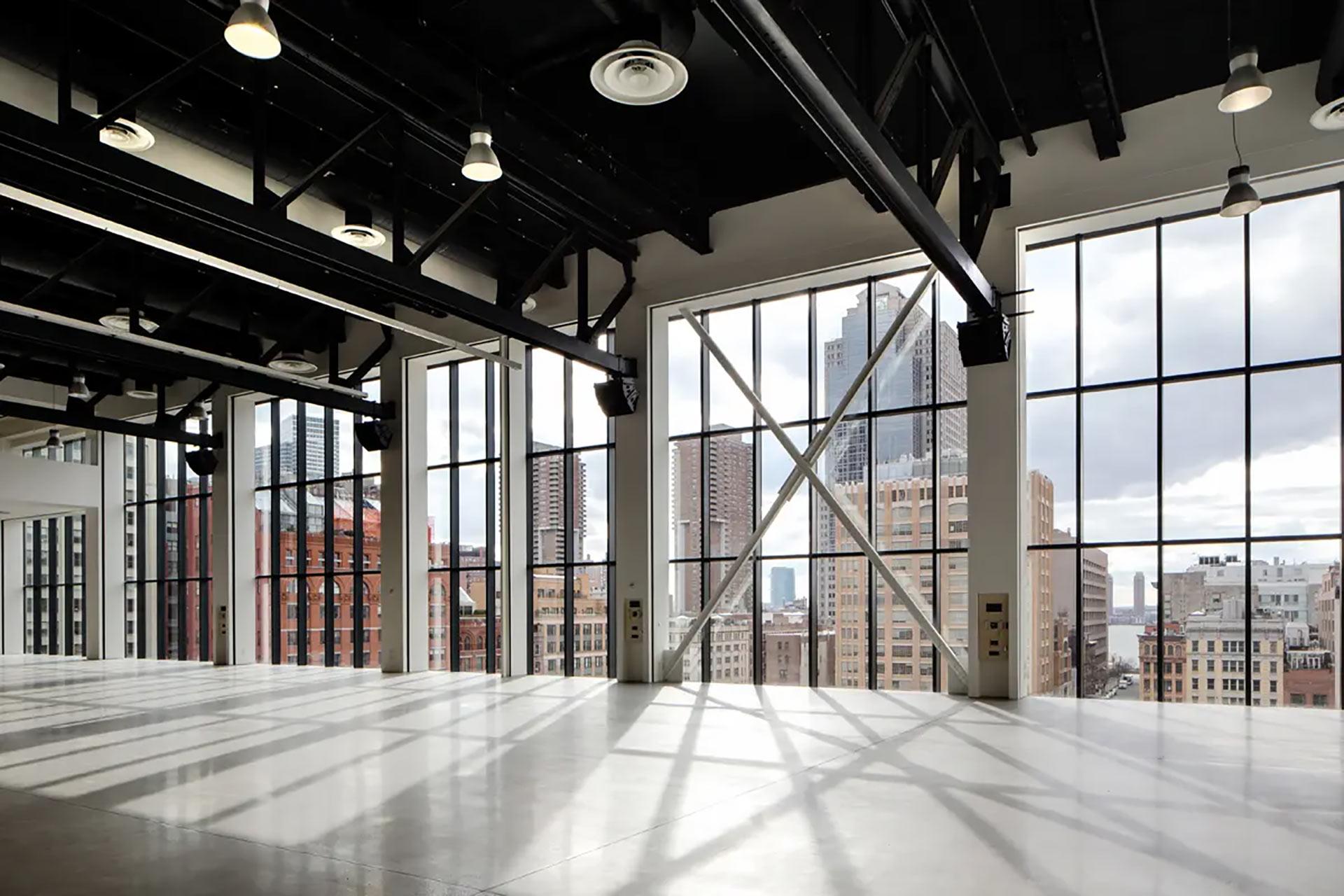A spacious, empty loft with large floor-to-ceiling industrial windows, black steel beams, and city skyscrapers visible outside in daylight. Sunlight casts geometric shadows on the polished concrete floor.