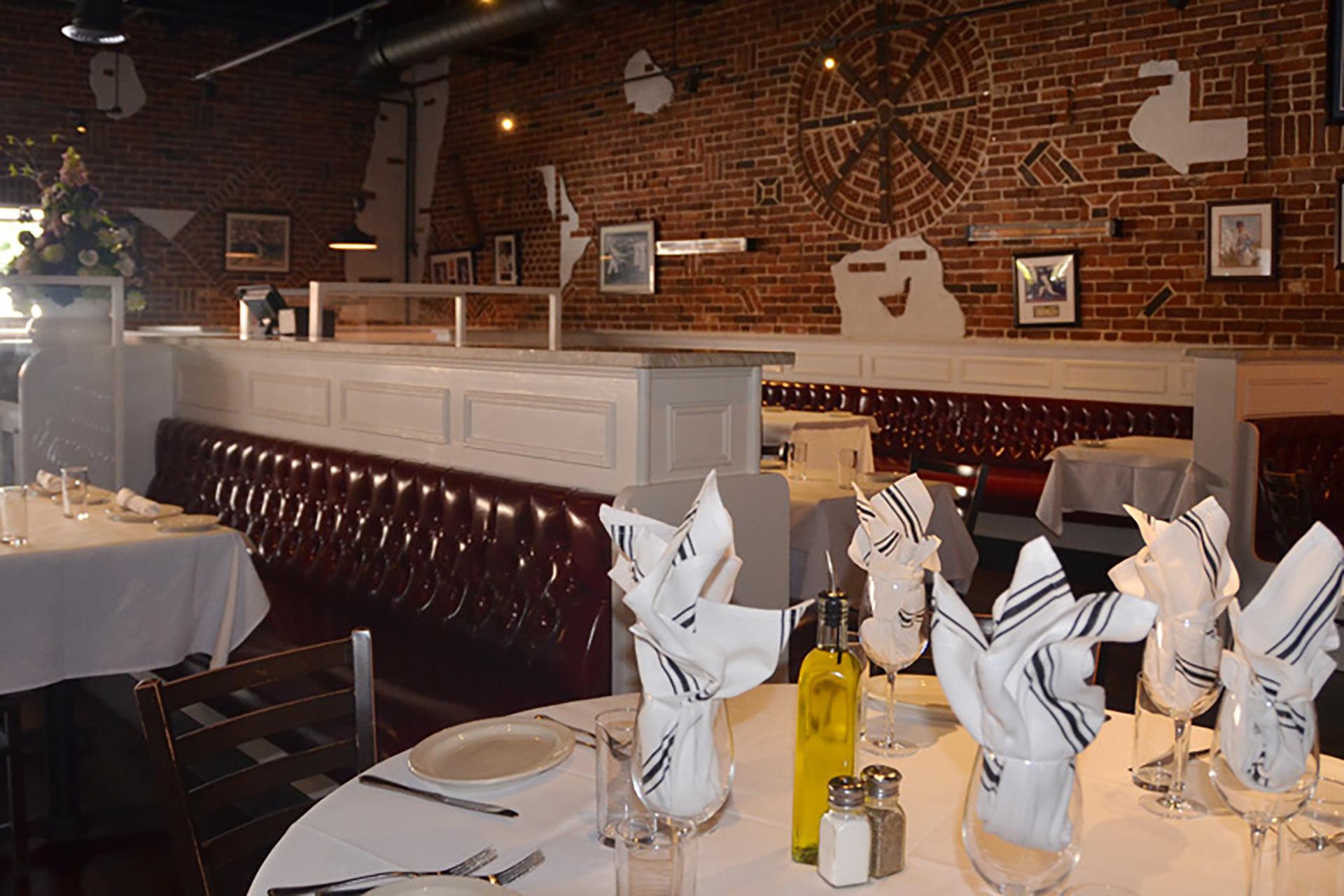 A restaurant dining area with white tablecloths, neatly folded napkins, glassware, and dark red leather booths. The exposed brick wall features white mural art and framed pictures. The atmosphere is cozy and inviting.