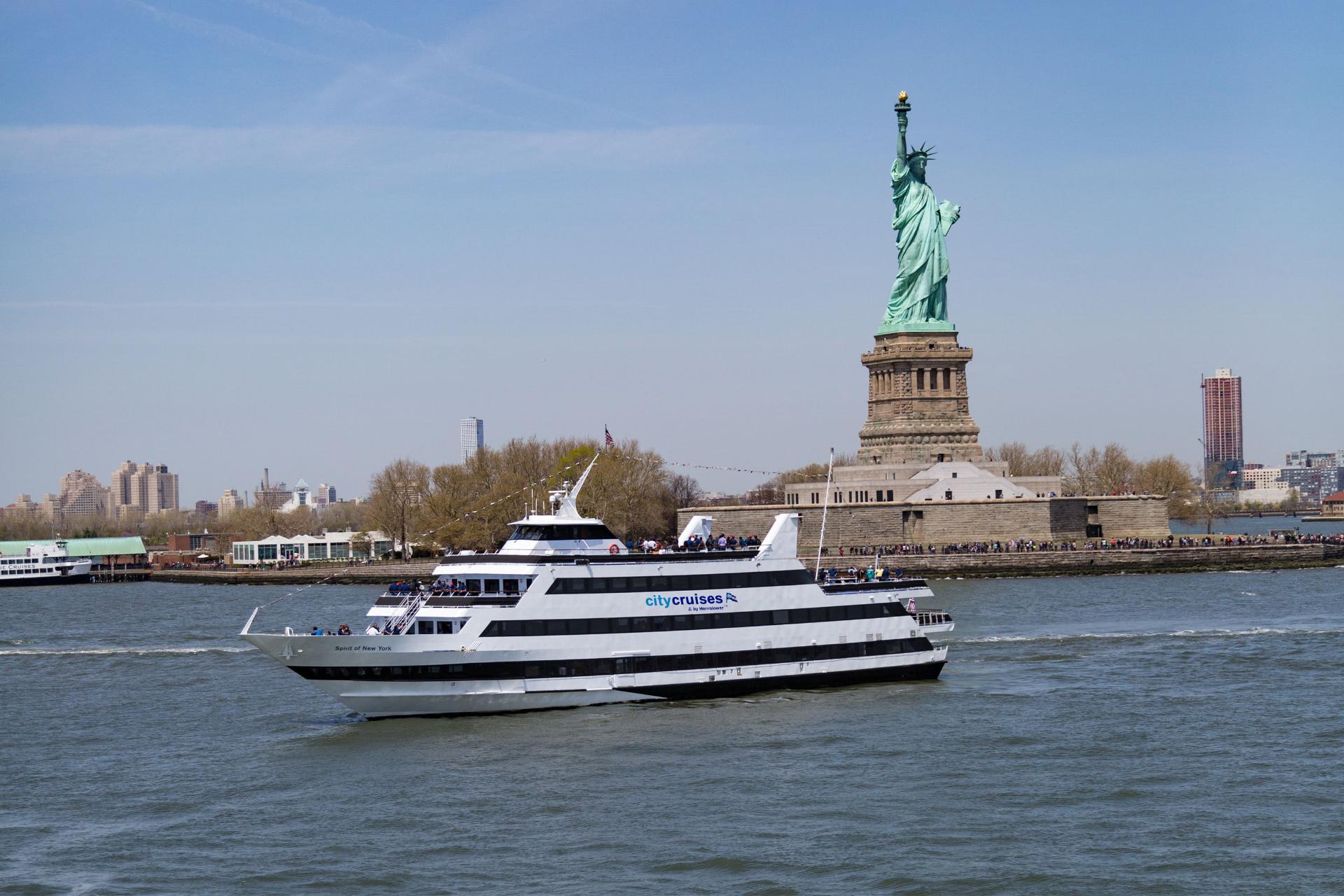 A white City Cruises ferry boat sails past the Statue of Liberty on a sunny day, with passengers on deck and the New York City skyline in the background.