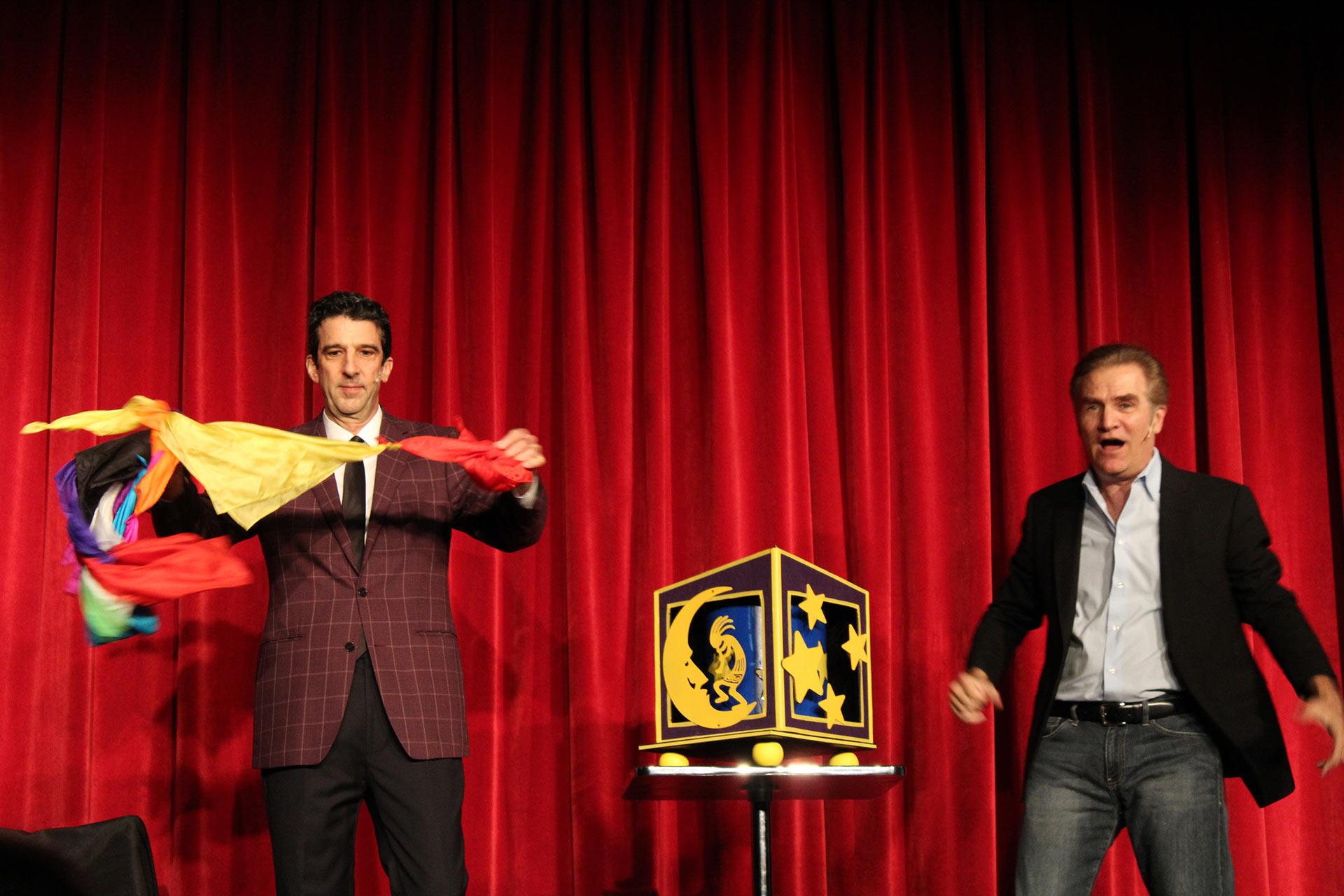 Two men perform a magic act on stage in front of closed red curtains. One man holds colorful scarves, and the other stands beside a decorated box on a small table. Both are dressed in suits and look engaged in the performance.