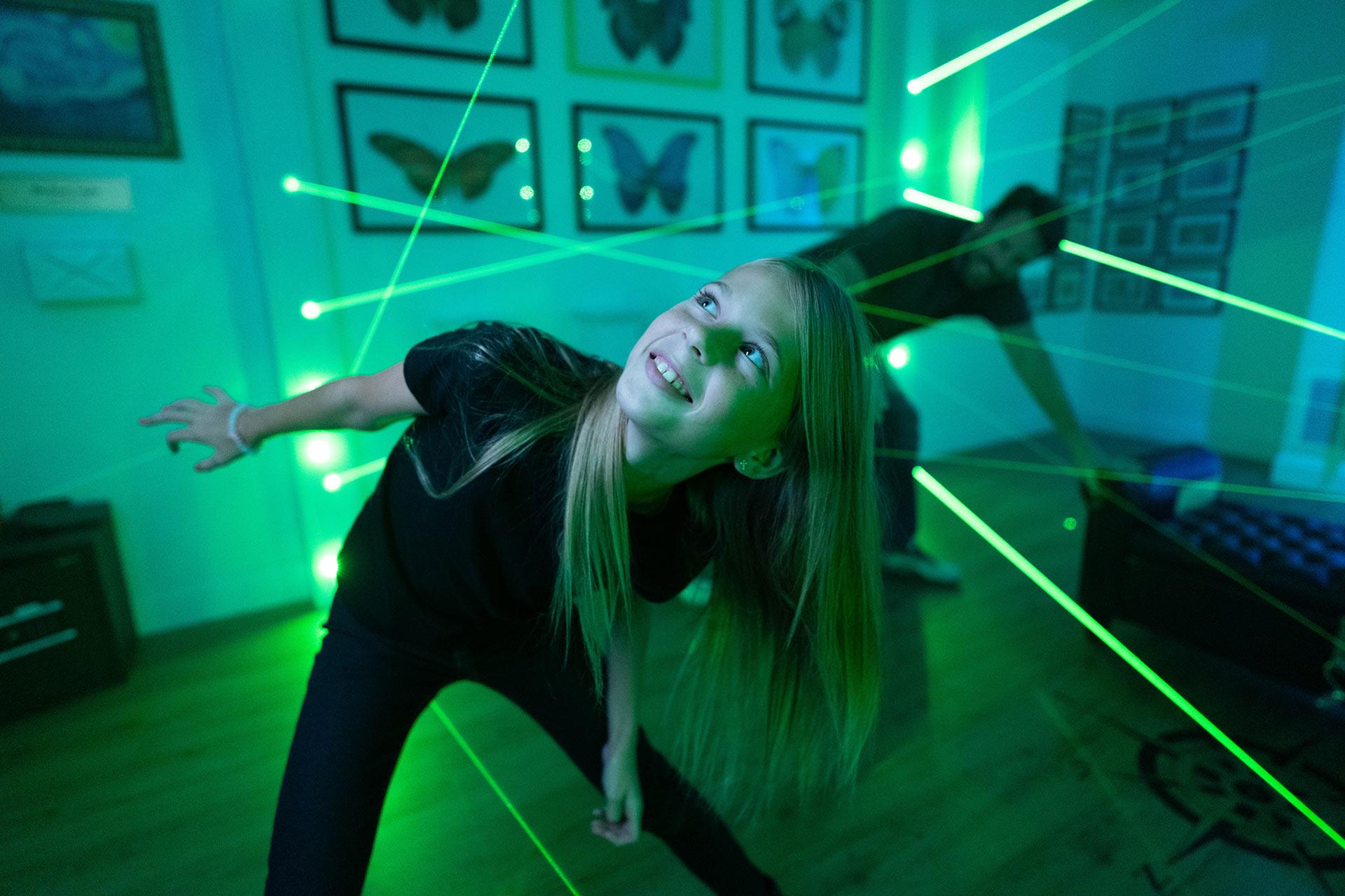 A smiling girl weaves through green laser beams in a room decorated with butterfly art, playing an interactive laser maze game.