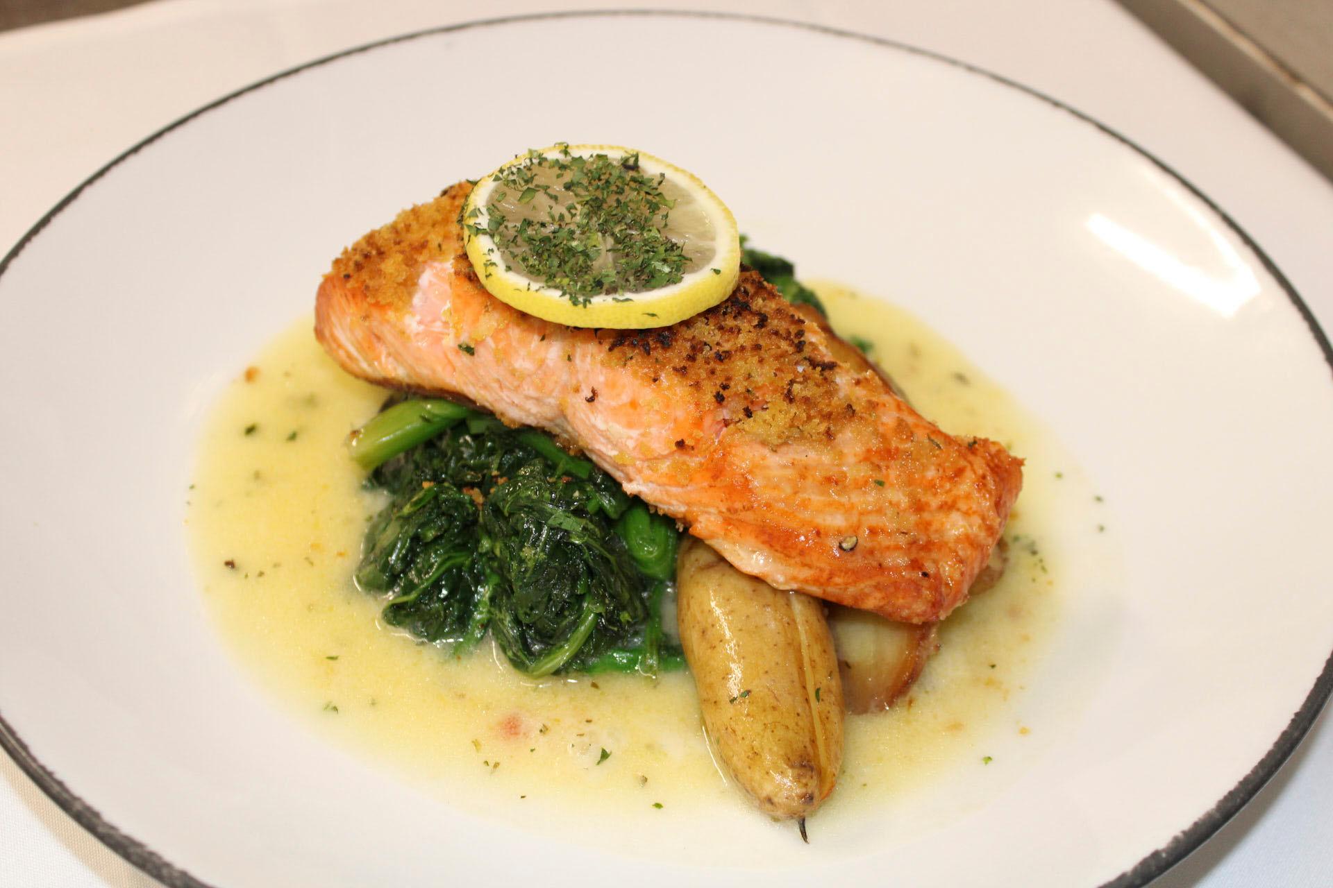 A plated dish featuring seared salmon topped with a lemon slice and herbs, served on a bed of sautéed greens and fingerling potatoes, all in a creamy sauce.