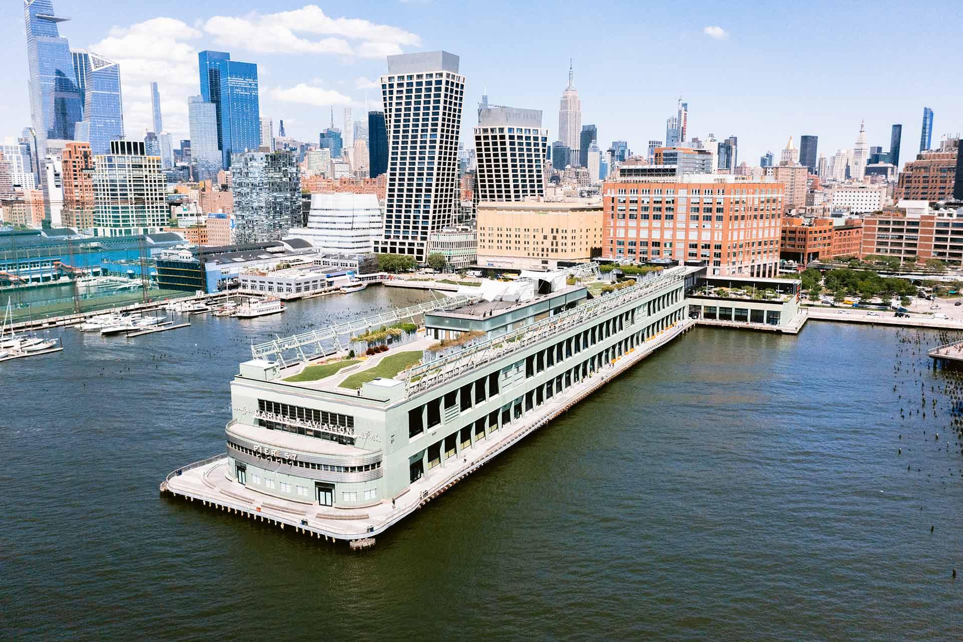 A large waterfront building with a rooftop garden extends into the river, surrounded by docks, with New York City skyscrapers and mid-rise buildings visible in the background under a blue sky.