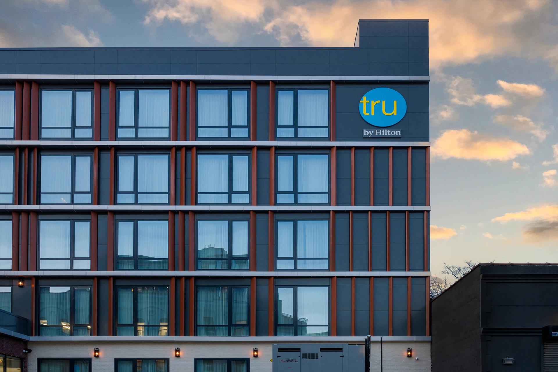 A modern hotel building with large windows and a blue and yellow "tru by Hilton" sign on the upper corner, set against a partly cloudy sky at sunset.