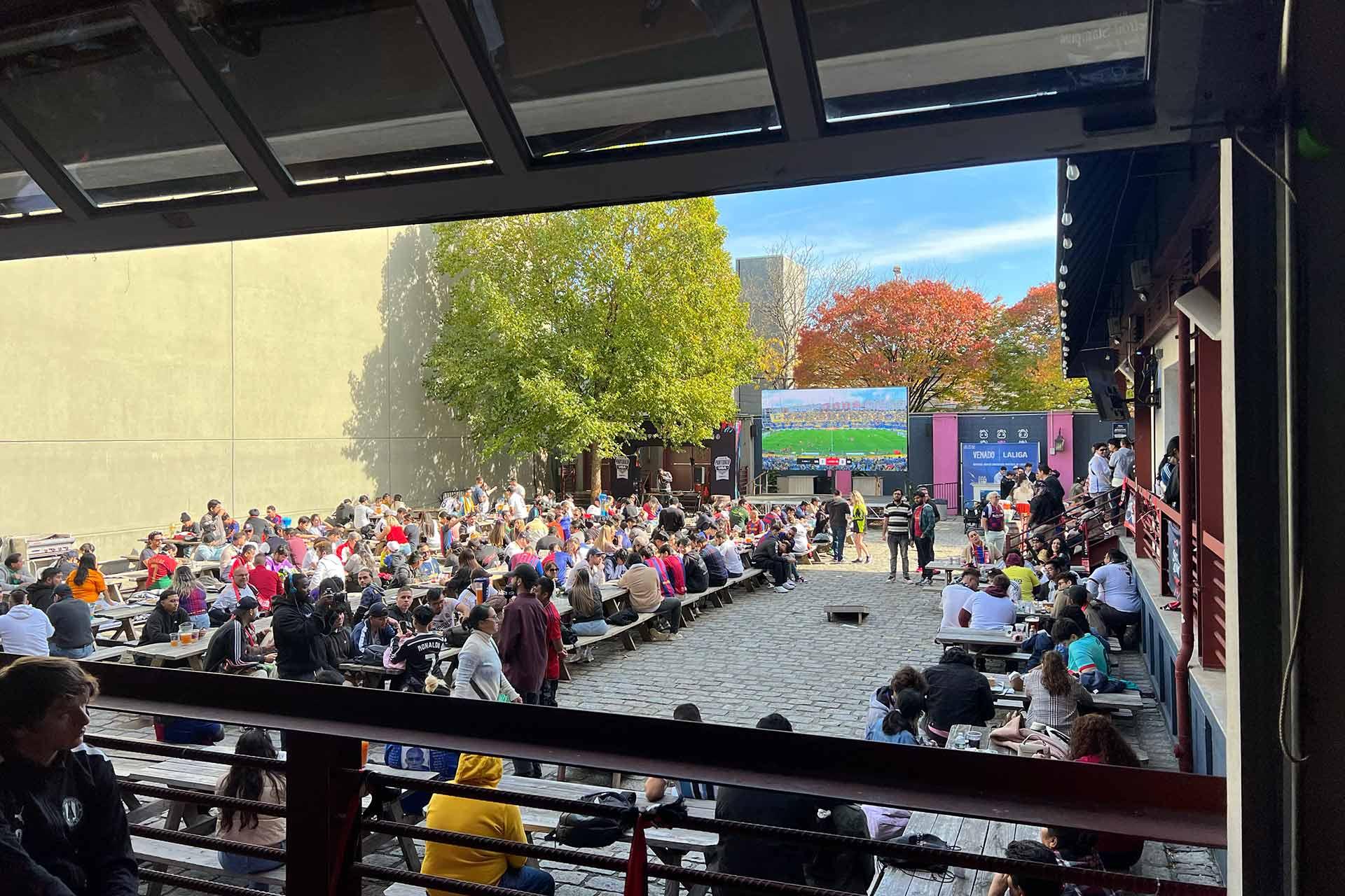 A large crowd sits at outdoor picnic tables watching a sporting event on a big screen in a courtyard, surrounded by trees with green and autumn-colored leaves. Some people are standing or walking by.