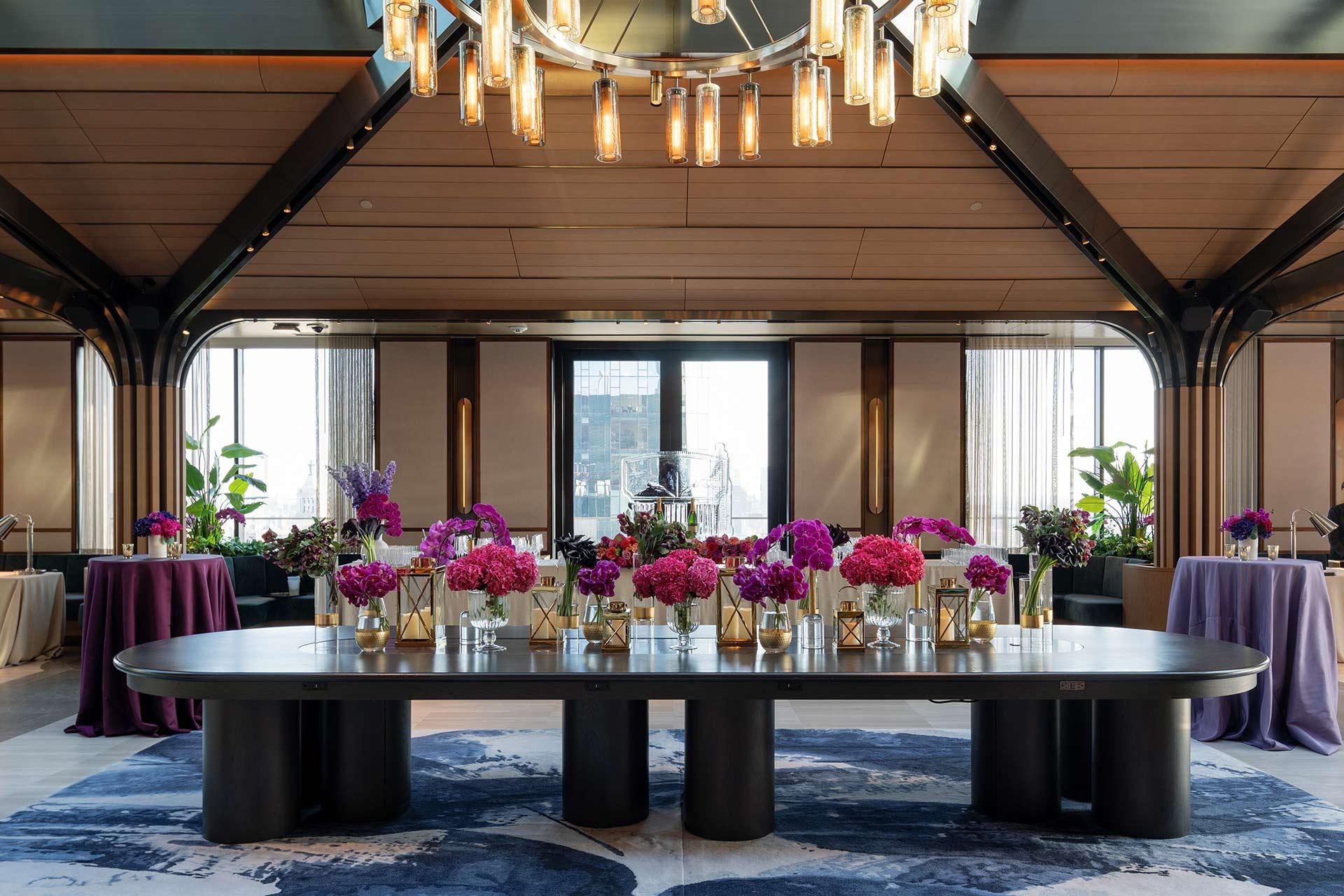 A modern, elegant room with a large black table decorated with vibrant pink and purple flower arrangements in gold vases. Tall windows, a chandelier, and additional tables with similar decor are visible in the background.