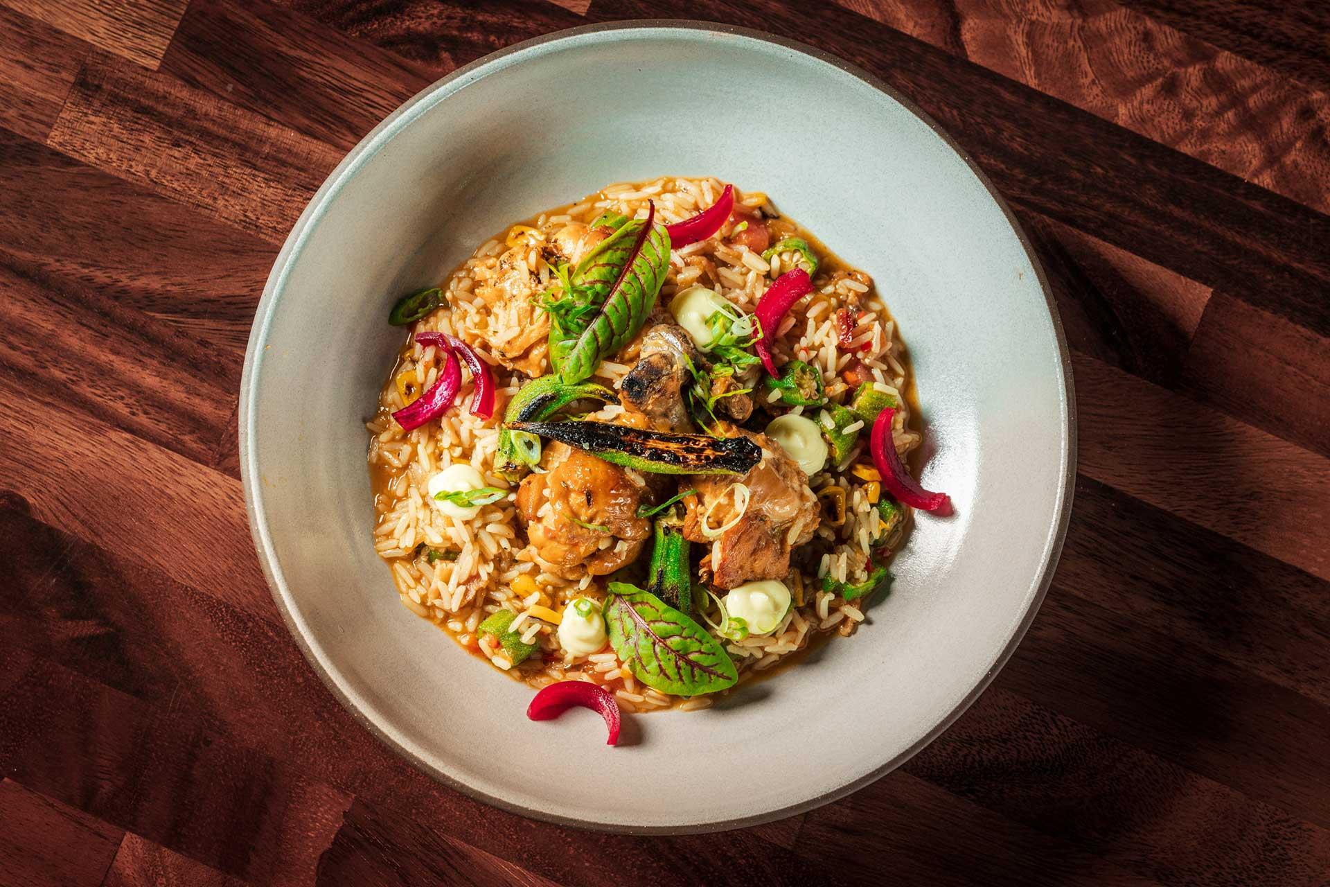 A bowl of rice topped with pieces of grilled meat, green herbs, red pickled onions, and dollops of creamy sauce, served on a white plate placed on a wooden surface.