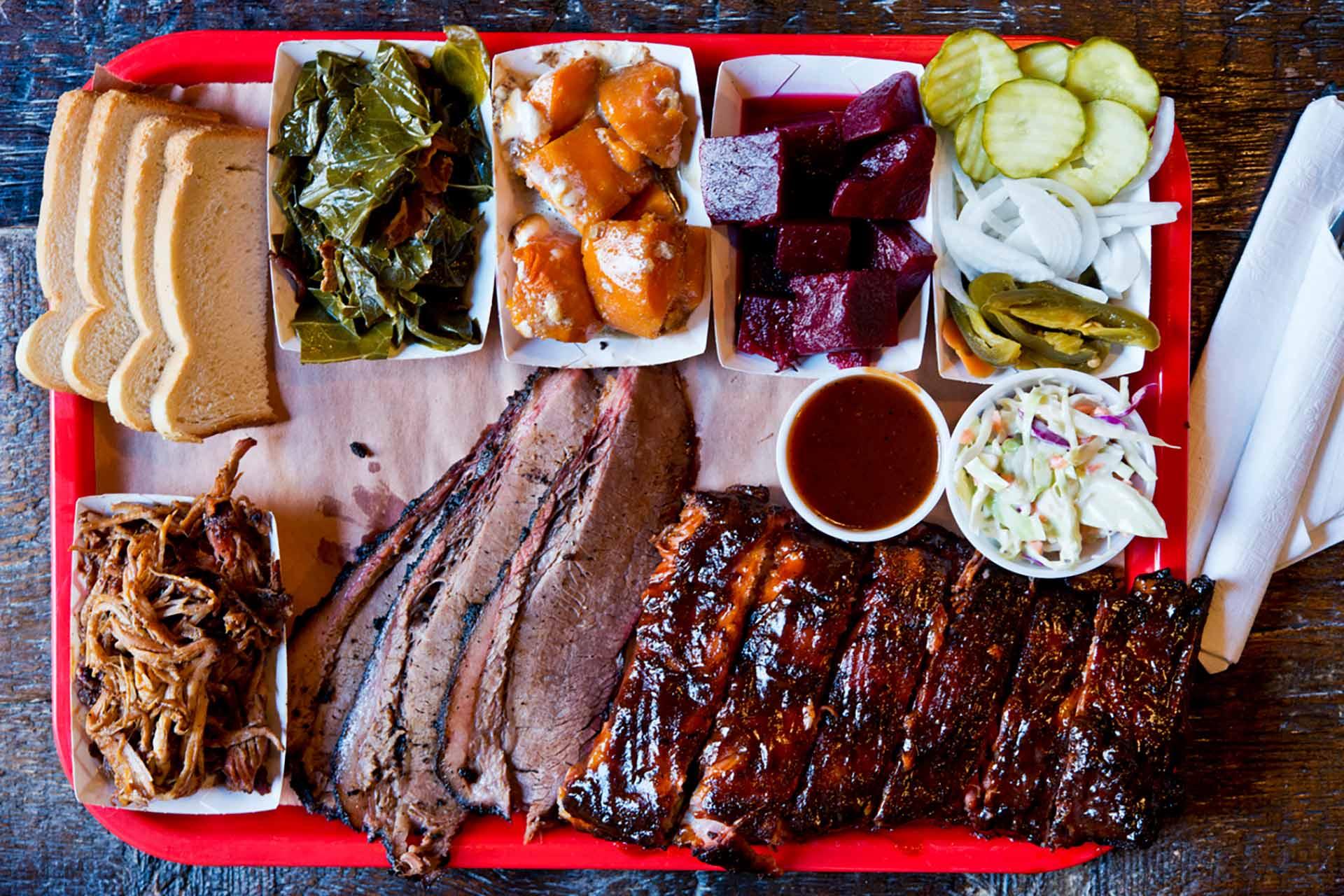 A tray of barbecue food including sliced brisket, ribs with sauce, pulled pork, sliced bread, collard greens, sweet potatoes, beets, pickles, onions, jalapeños, coleslaw, and a cup of barbecue sauce.