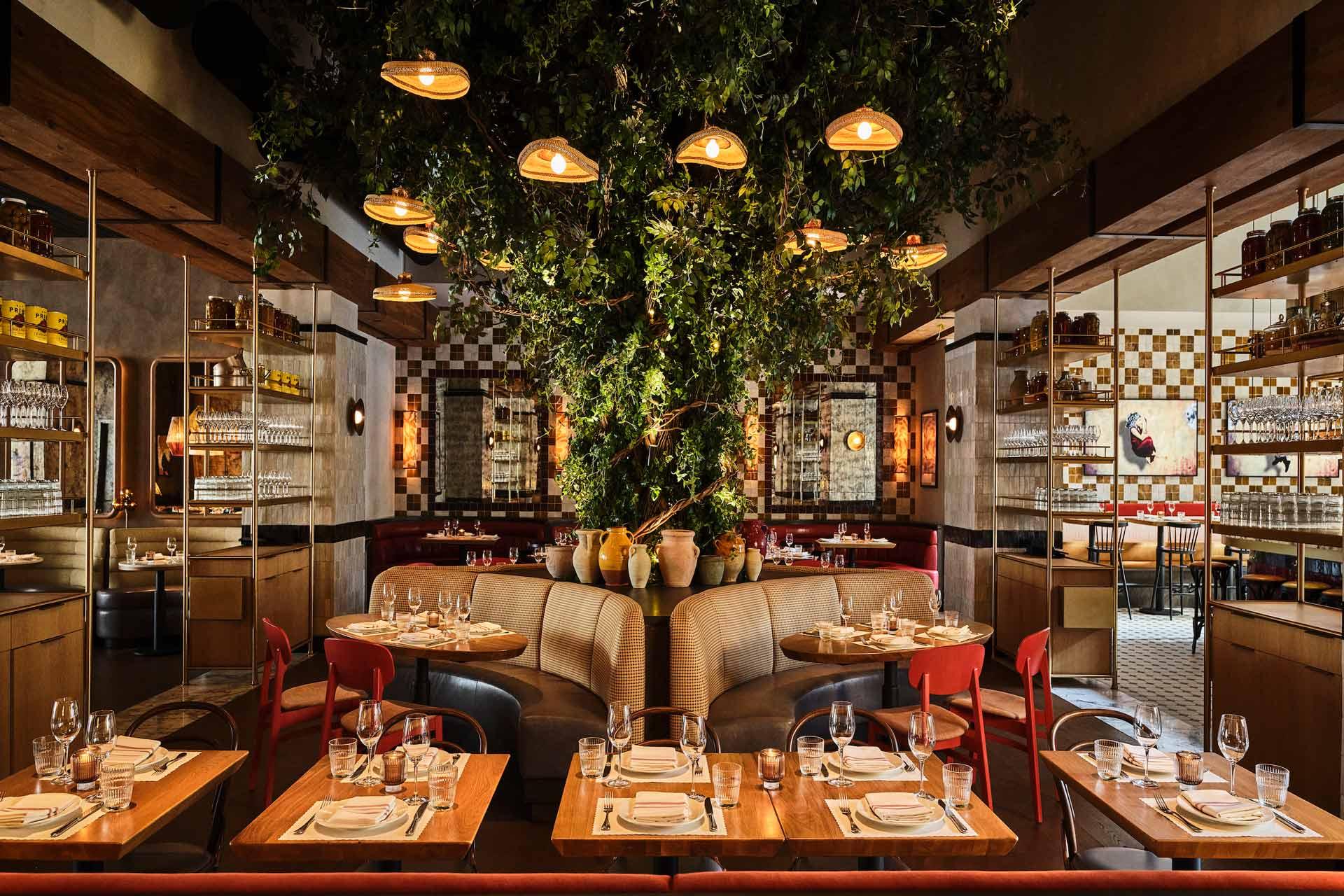 Elegant restaurant interior with wooden tables set for dining, cushioned round booth seating, red chairs, shelves with glassware, and a large tree with hanging lights as a centerpiece. Warm, ambient lighting throughout.