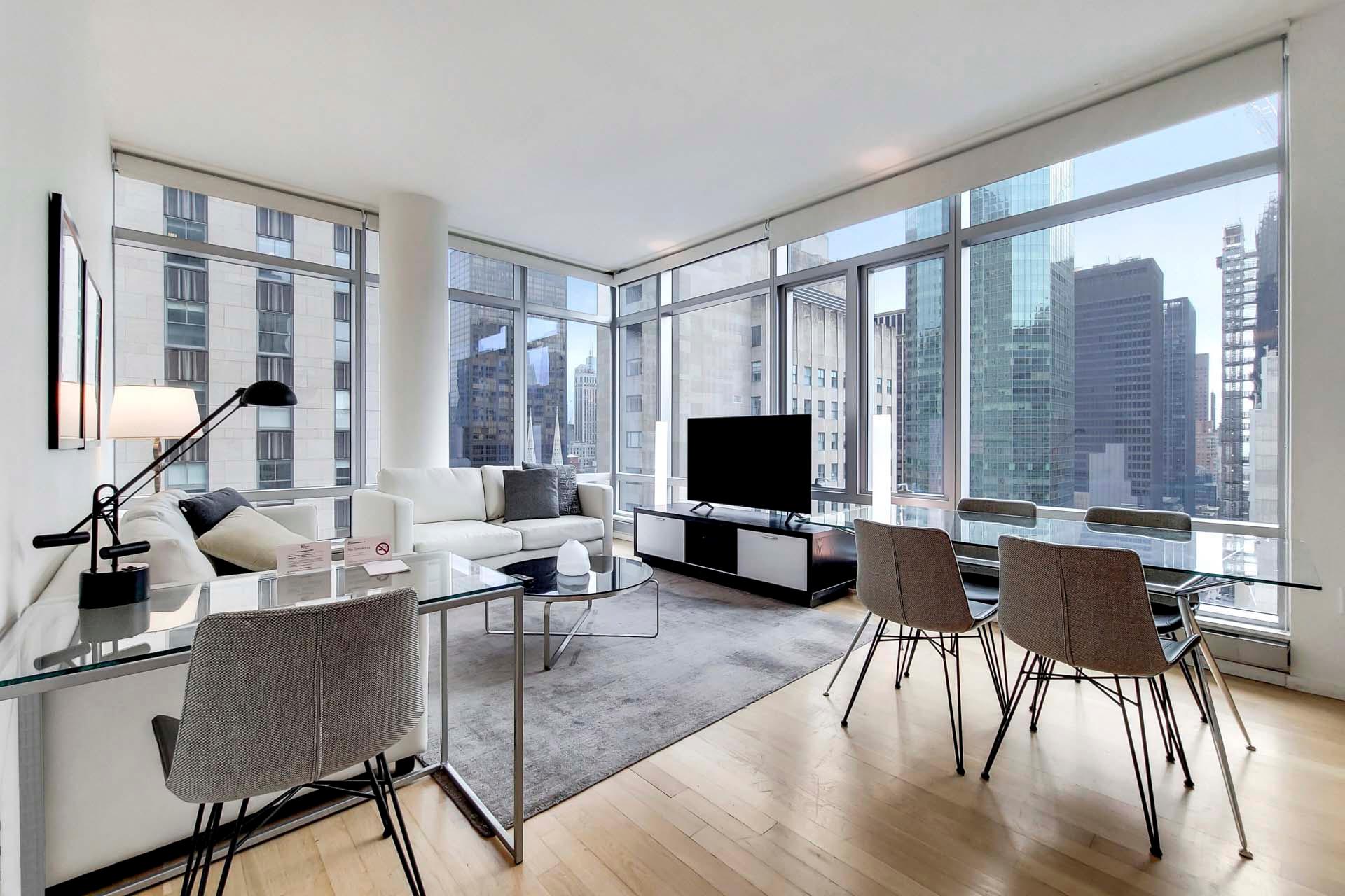 Modern living room with large windows showcasing a cityscape view. It features a white sofa, TV, glass dining table with gray chairs, and a work desk with a lamp. The room has a light wood floor and minimalistic decor.
