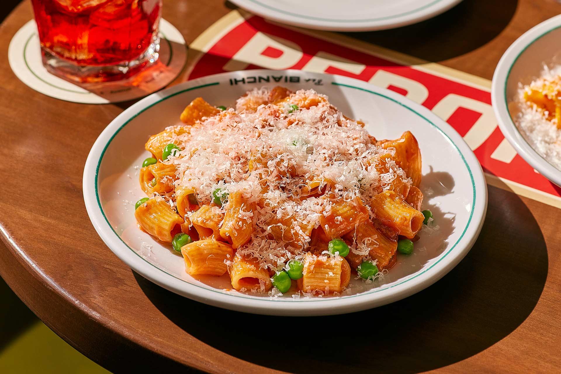 A plate of rigatoni pasta in tomato sauce topped with grated cheese and green peas sits on a wooden table next to a red drink in a glass.
