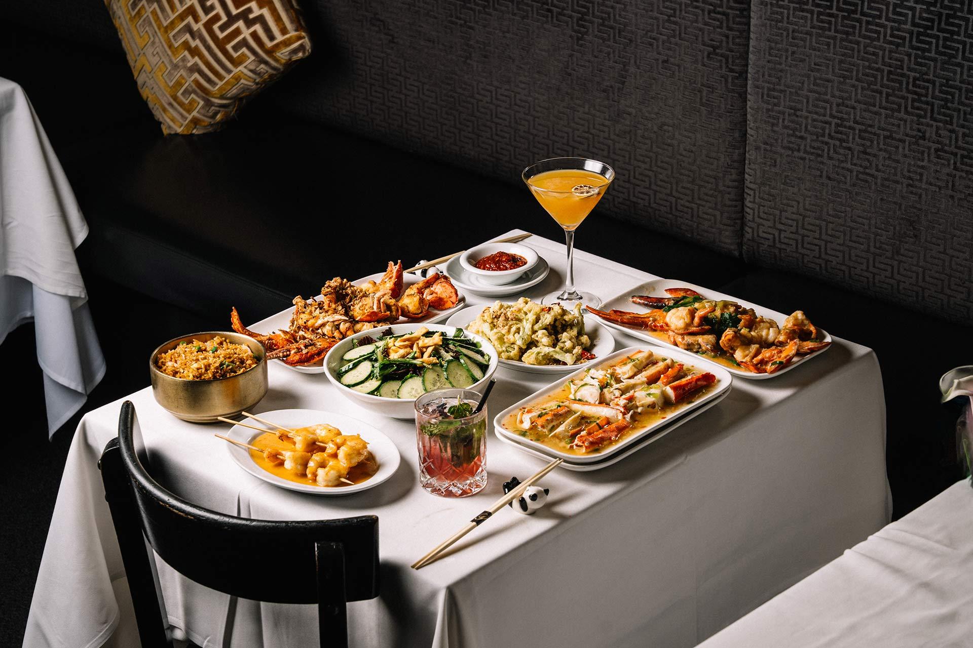 A table with white tablecloth set with various seafood dishes, vegetables, fried rice, sauces, and two cocktails, in a modern restaurant with cushioned seating and decorative pillows.