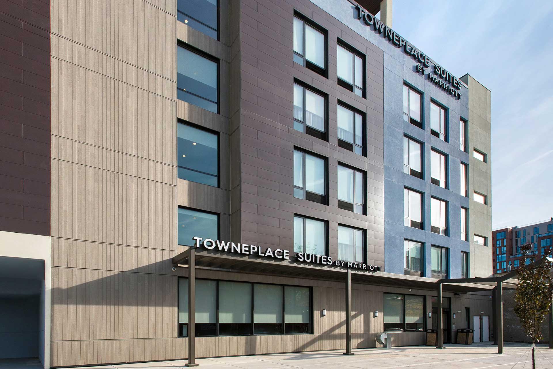 The exterior of a modern TownePlace Suites by Marriott hotel, featuring large windows, a flat roof, and a covered entrance area with the hotel name displayed above the entrance.
