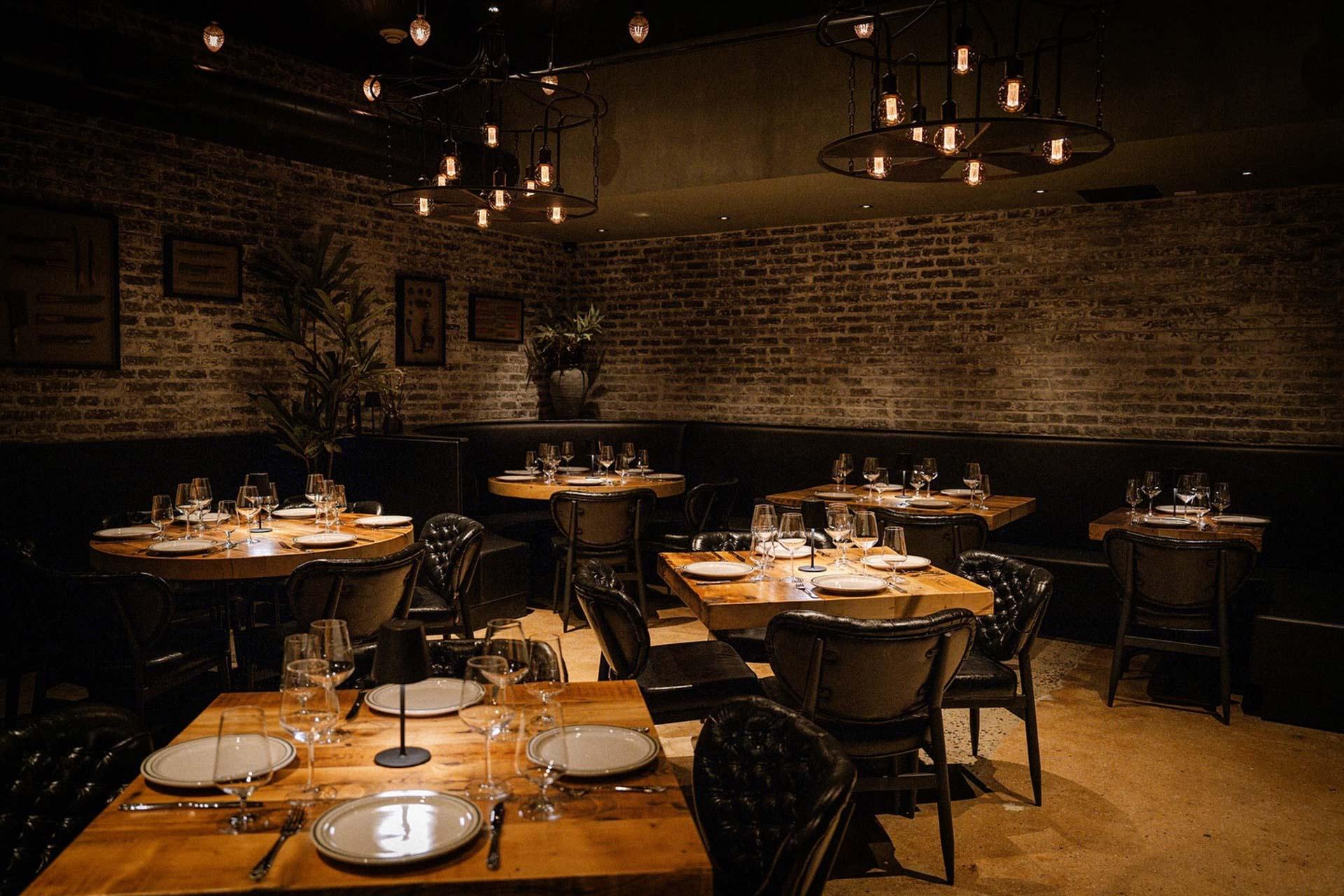 A cozy, dimly lit restaurant with exposed brick walls, wooden tables set with plates, cutlery, and glasses, black cushioned chairs, decorative plants, and hanging industrial-style light fixtures.