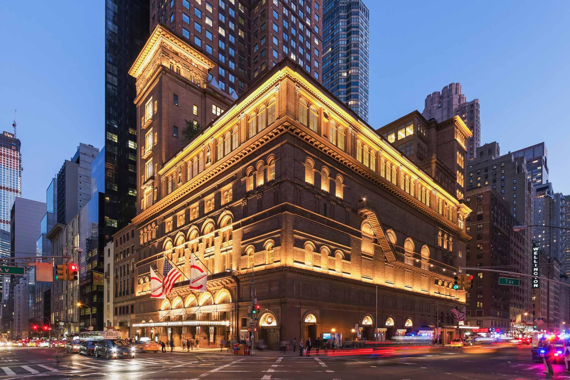 A grand, historic building with warm, illuminated lights highlighting its intricate architecture at dusk. American flags hang by the entrance. Surrounding traffic and city lights create a vibrant urban scene.