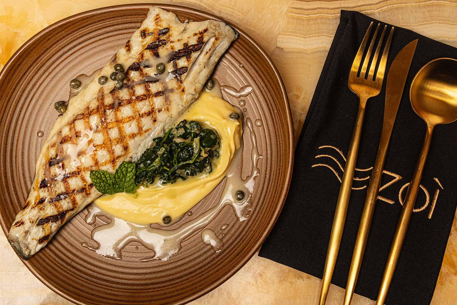 Grilled fish fillet with capers on top of creamy mashed potatoes and sautéed greens, served on a brown plate beside a black napkin with gold cutlery and "ZOI" logo.
