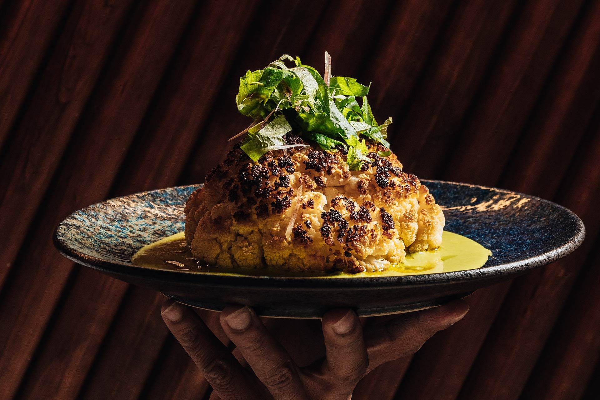 A hand holds a dark plate with a whole roasted cauliflower topped with fresh greens, sitting in a pool of yellow sauce, against a dark, textured background.