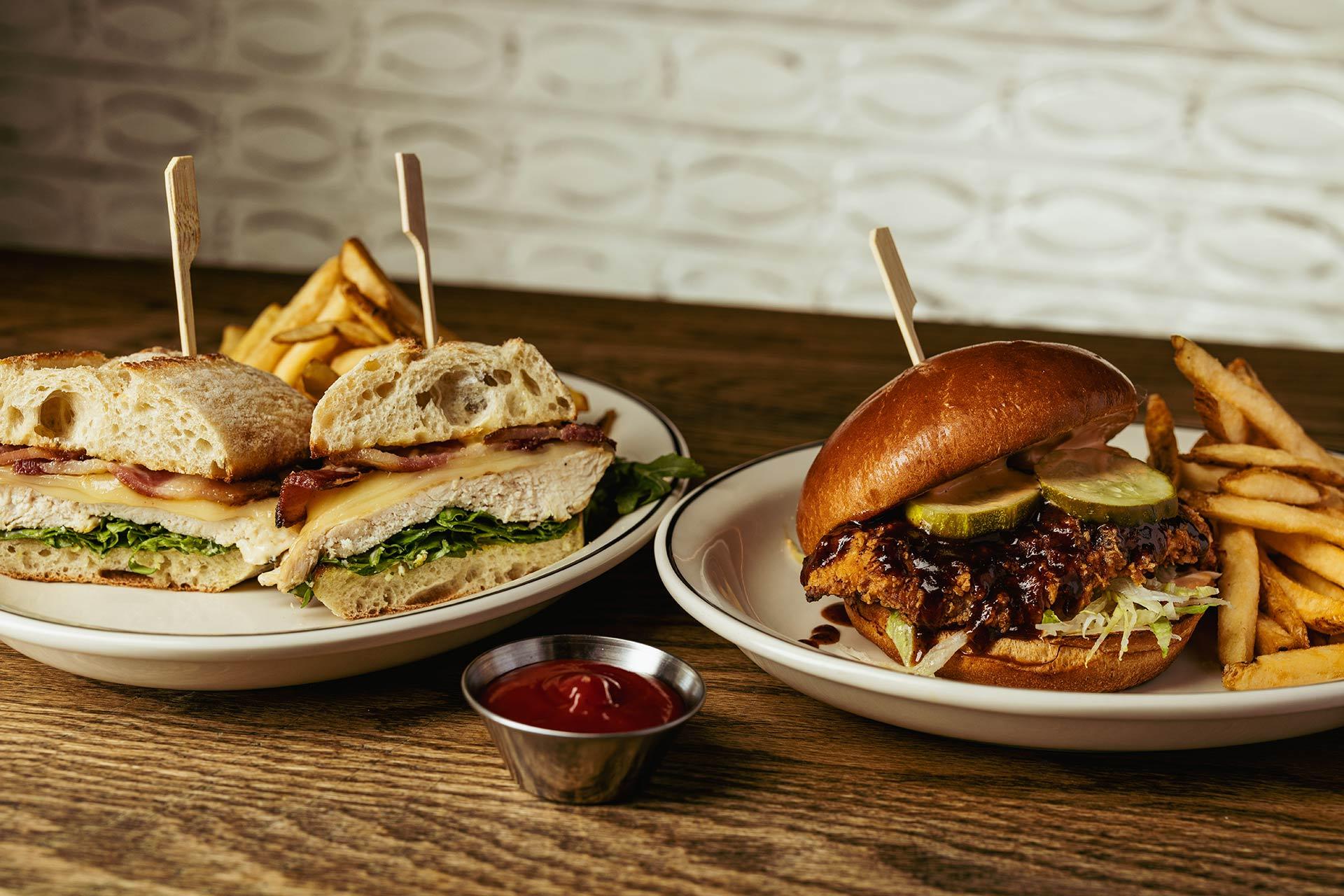 Two plates on a wooden table: one with a halved chicken sandwich, lettuce, bacon, cheese, and fries; the other with a barbecue chicken sandwich with pickles, fries, and a small metal cup of ketchup nearby.