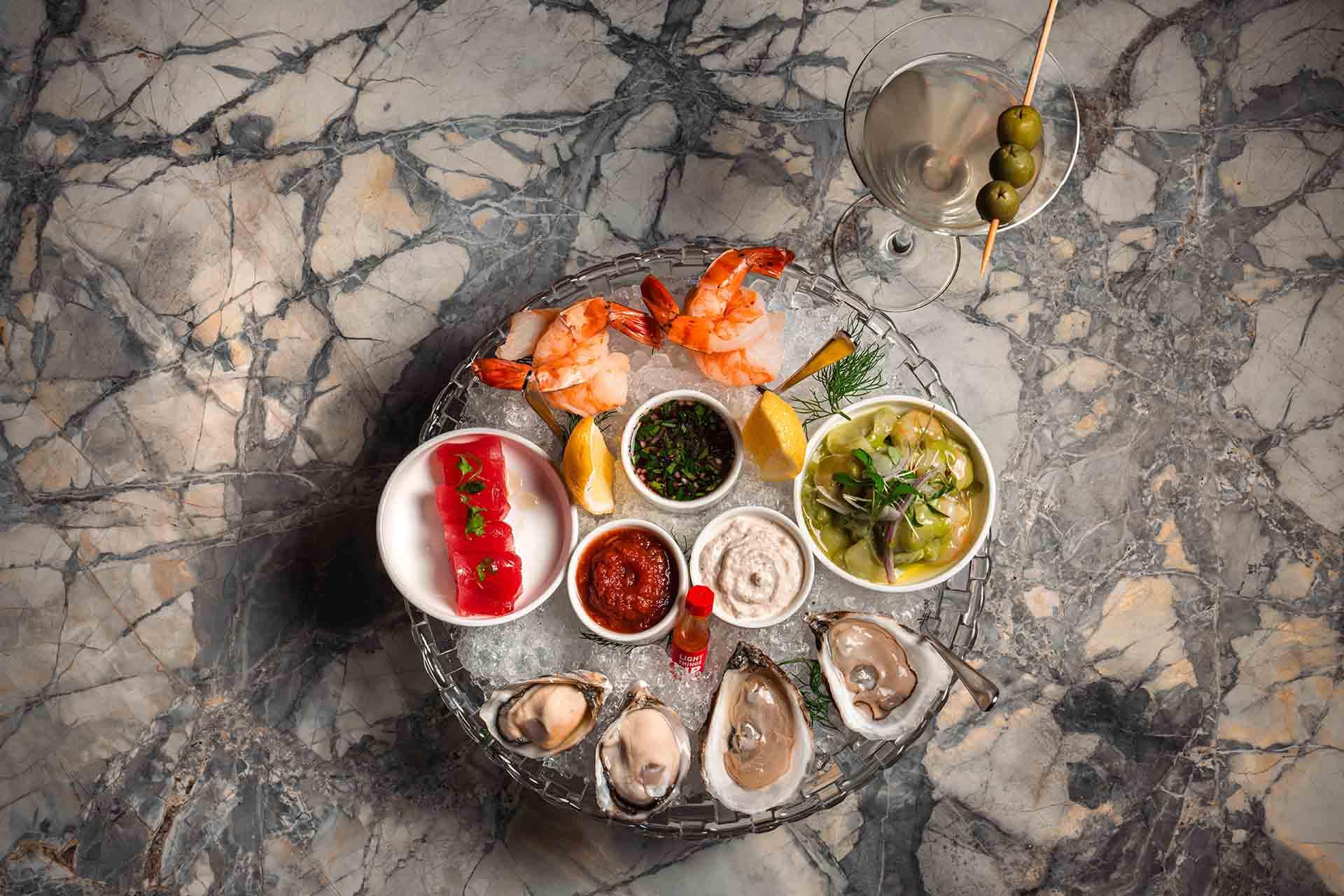 A marble table holds a seafood platter with oysters, shrimp, lemon wedges, sauces, tuna tartare, ceviche, and a martini garnished with green olives.