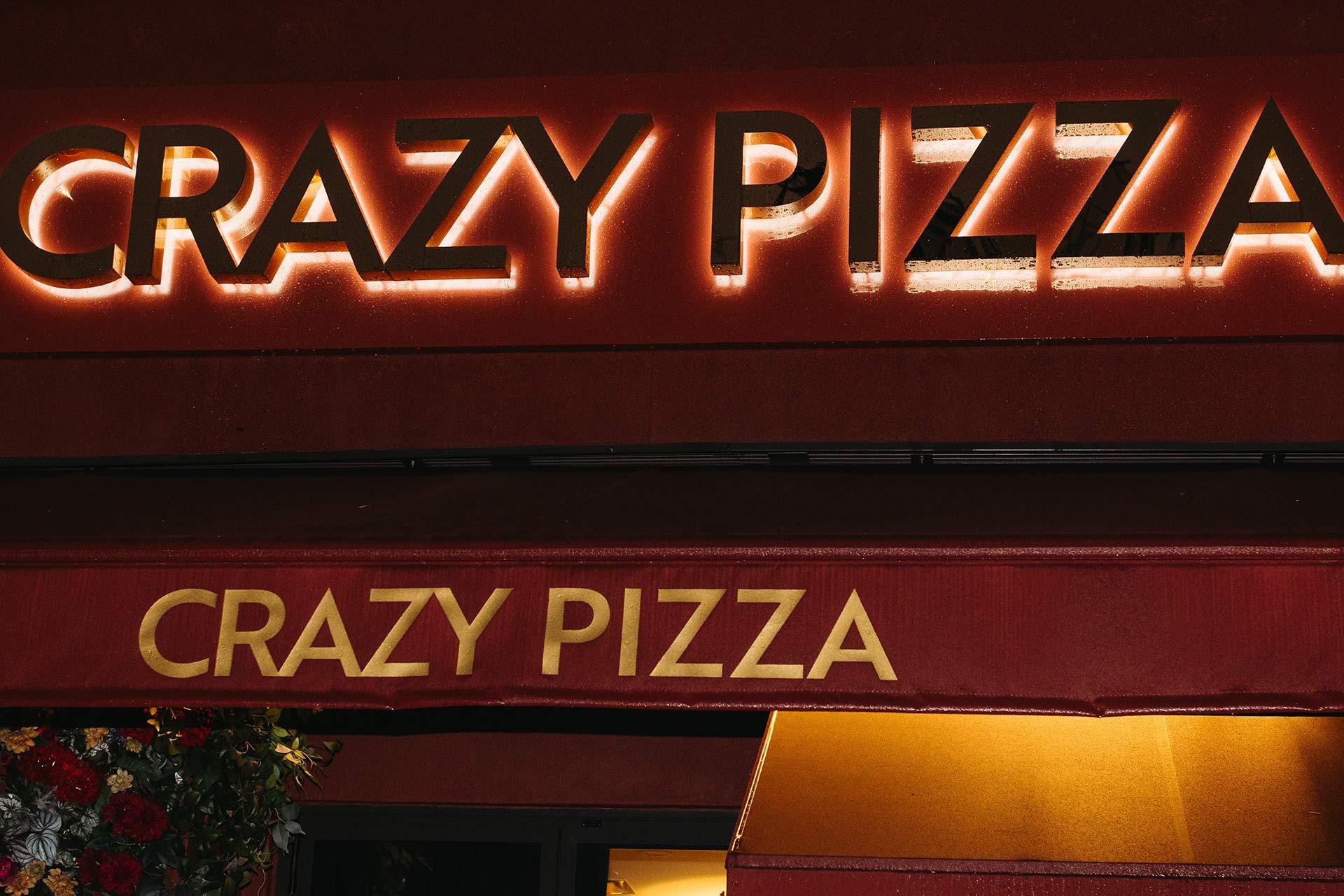 A restaurant sign and awning both display the name "CRAZY PIZZA" in bold, illuminated letters against a red background, with some flowers visible on the left side.
