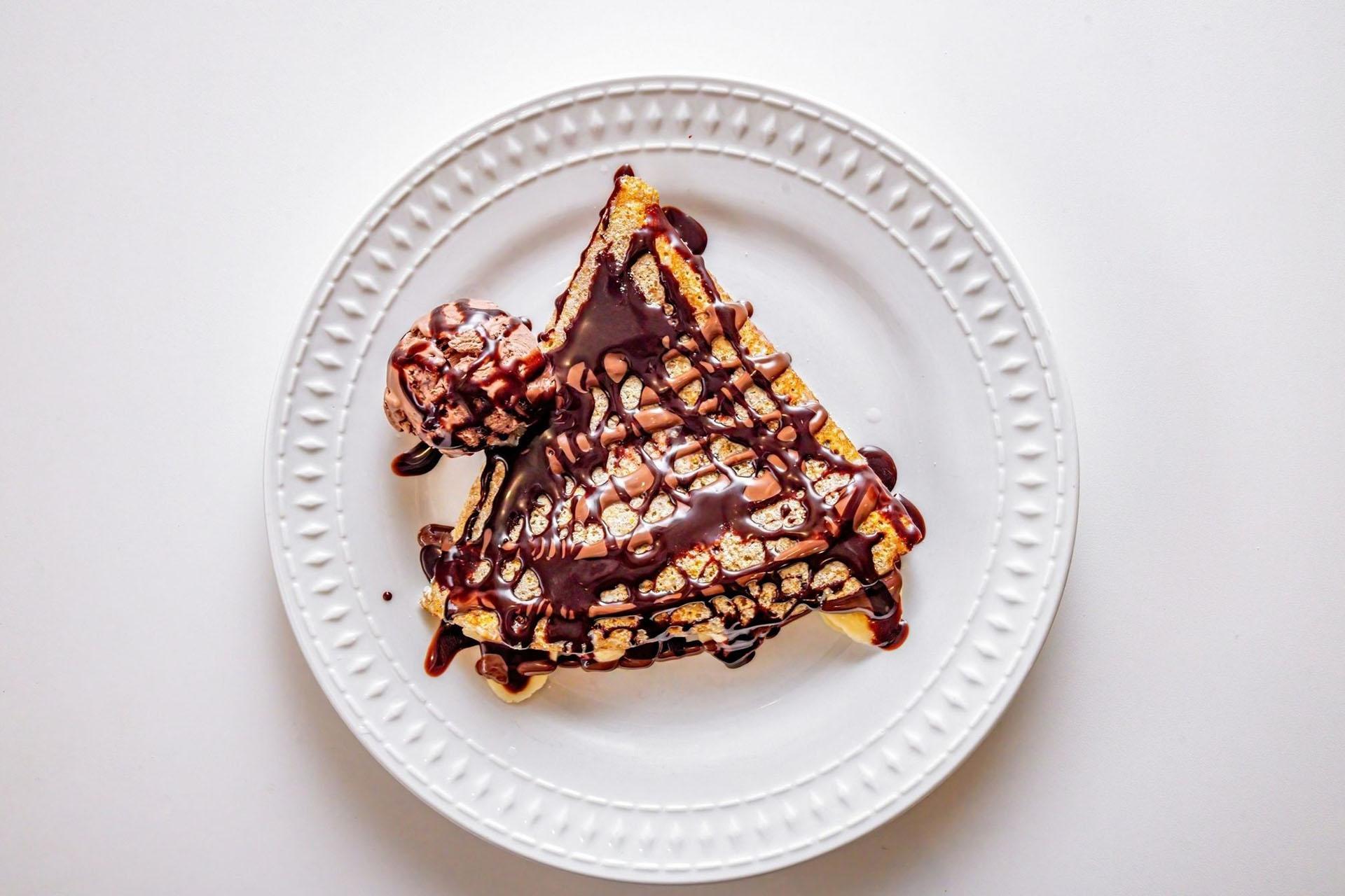 A triangular waffle topped with chocolate sauce and a scoop of chocolate ice cream sits on a white plate against a white background.