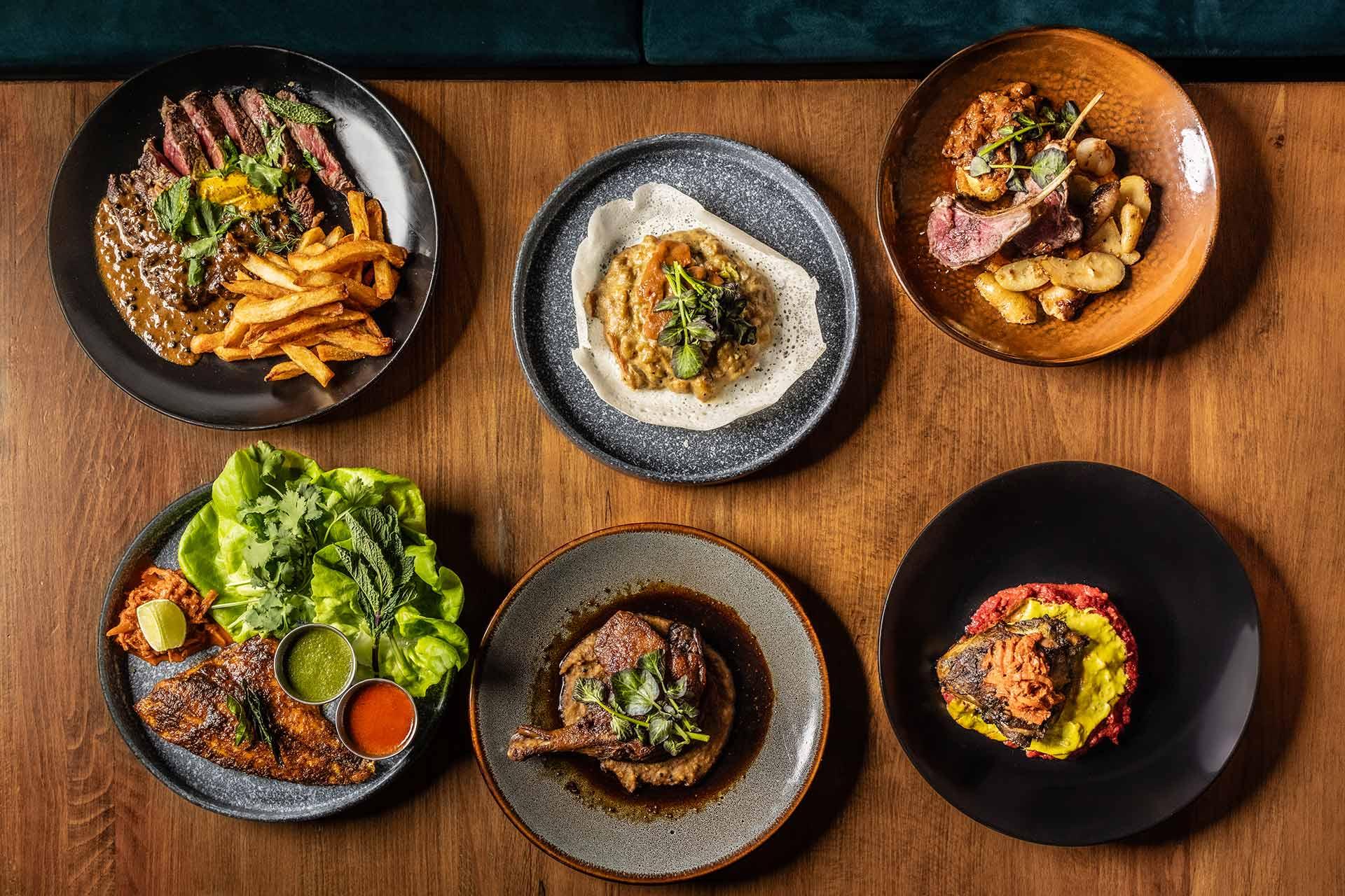 Six plates of assorted gourmet dishes, including steak with fries, grilled chicken, roasted meat with potatoes, a vegetable-topped dip, and dishes with colorful sauces, all arranged on a wooden table.