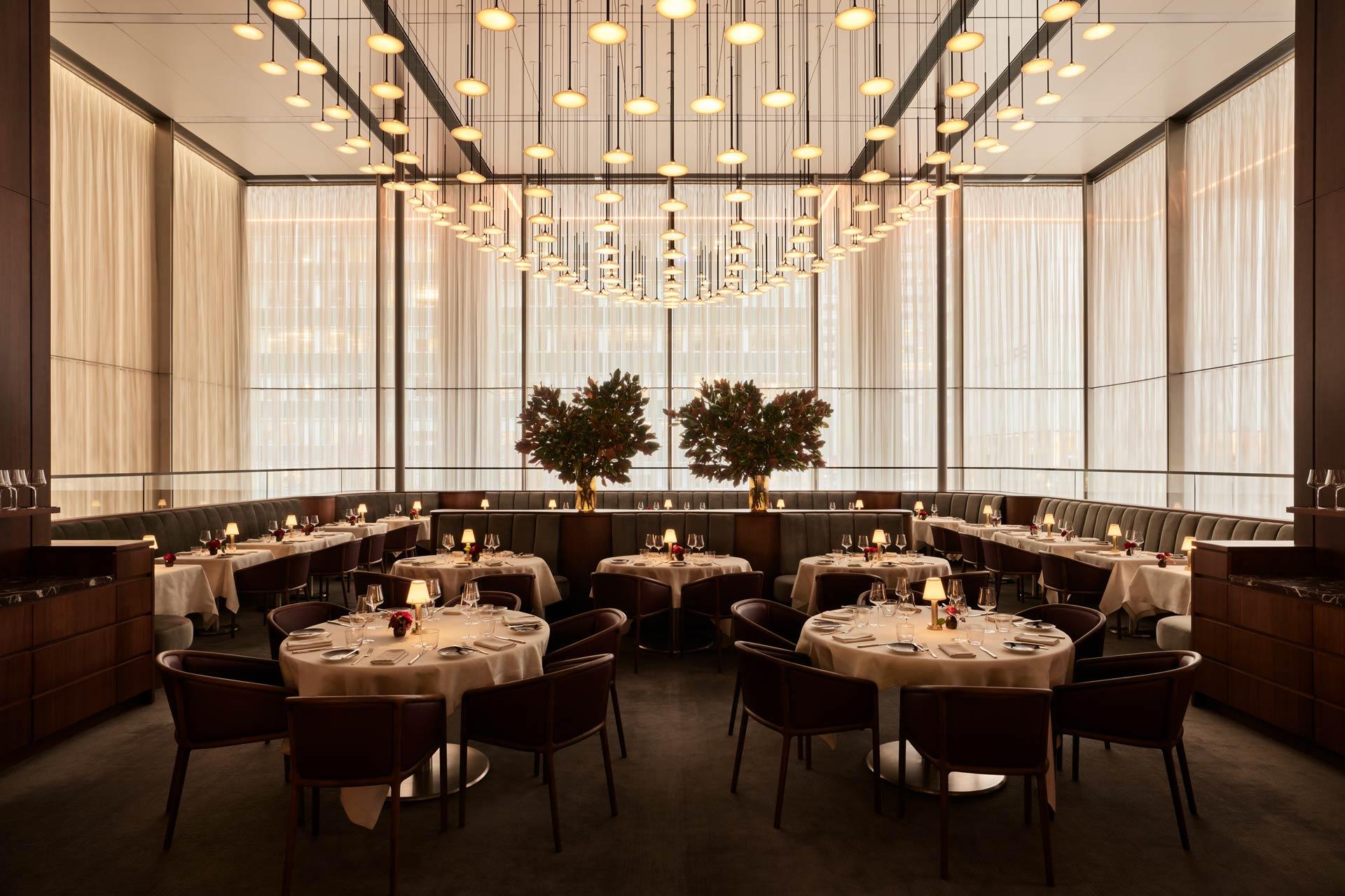 Elegant, modern restaurant dining room with round tables set for guests, leather chairs, warm lighting, tall windows with sheer curtains, and two large potted plants as a central feature.