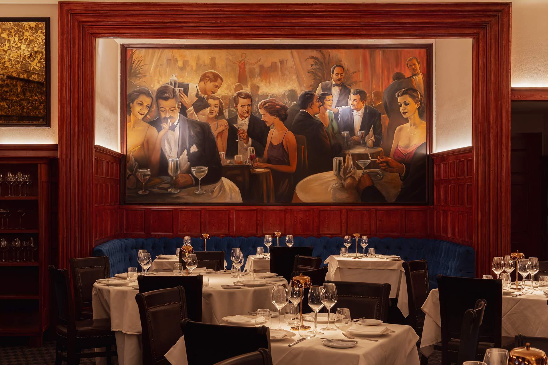 Elegant restaurant interior with white tablecloths, set tables, and dark wooden chairs. A large, colorful mural above blue banquette seating depicts a lively, glamorous dinner party scene from the early 20th century.