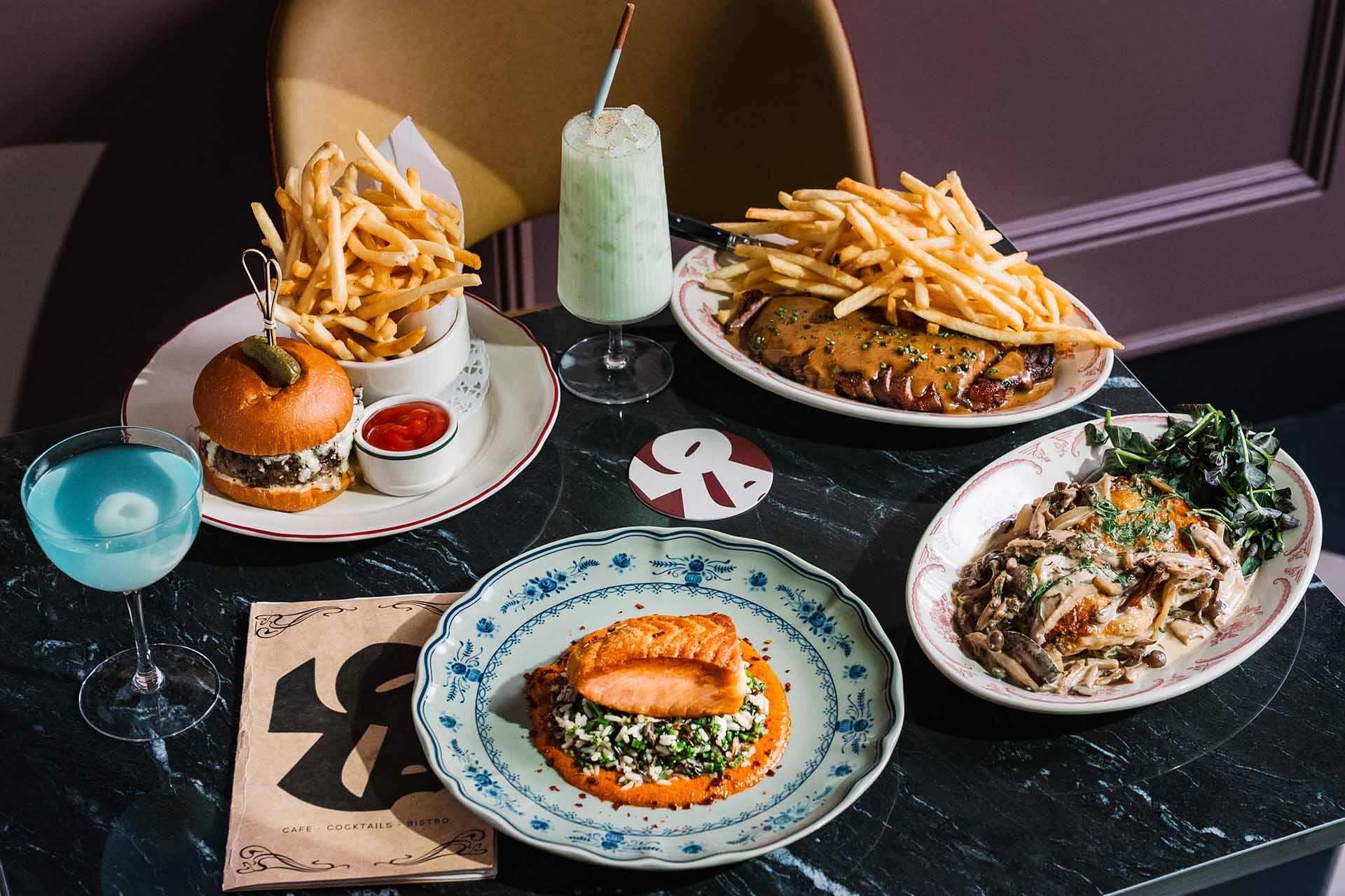A table set with a burger and fries, ribs with fries, pasta, grilled salmon with vegetables, a blue cocktail, a green milkshake, and a menu on a dark marble surface.