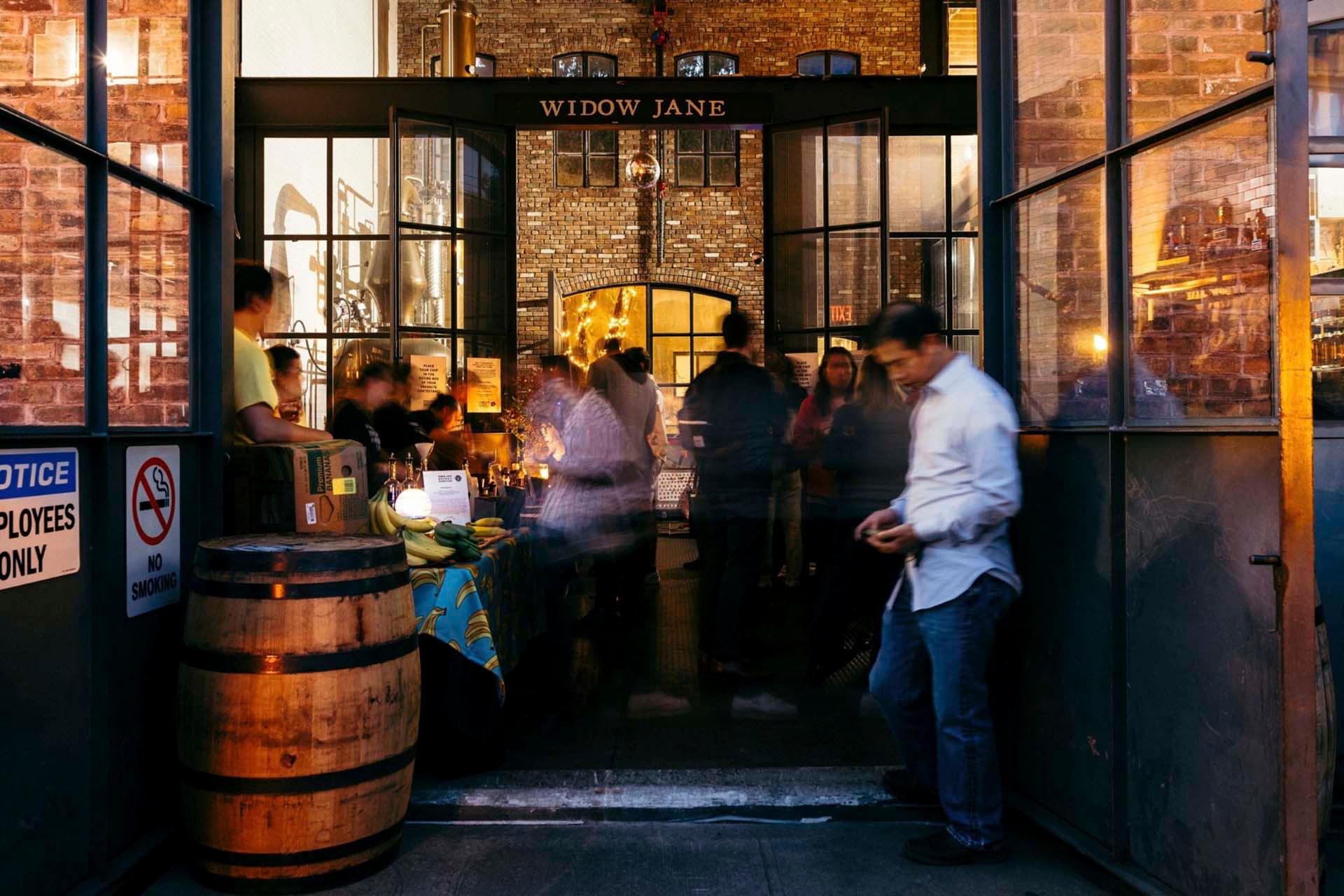 A group of people gather inside an open brick building labeled "Widow Jane," with a wooden barrel by the entrance and a man standing outside. Warm lighting glows from inside, creating a lively, welcoming atmosphere.