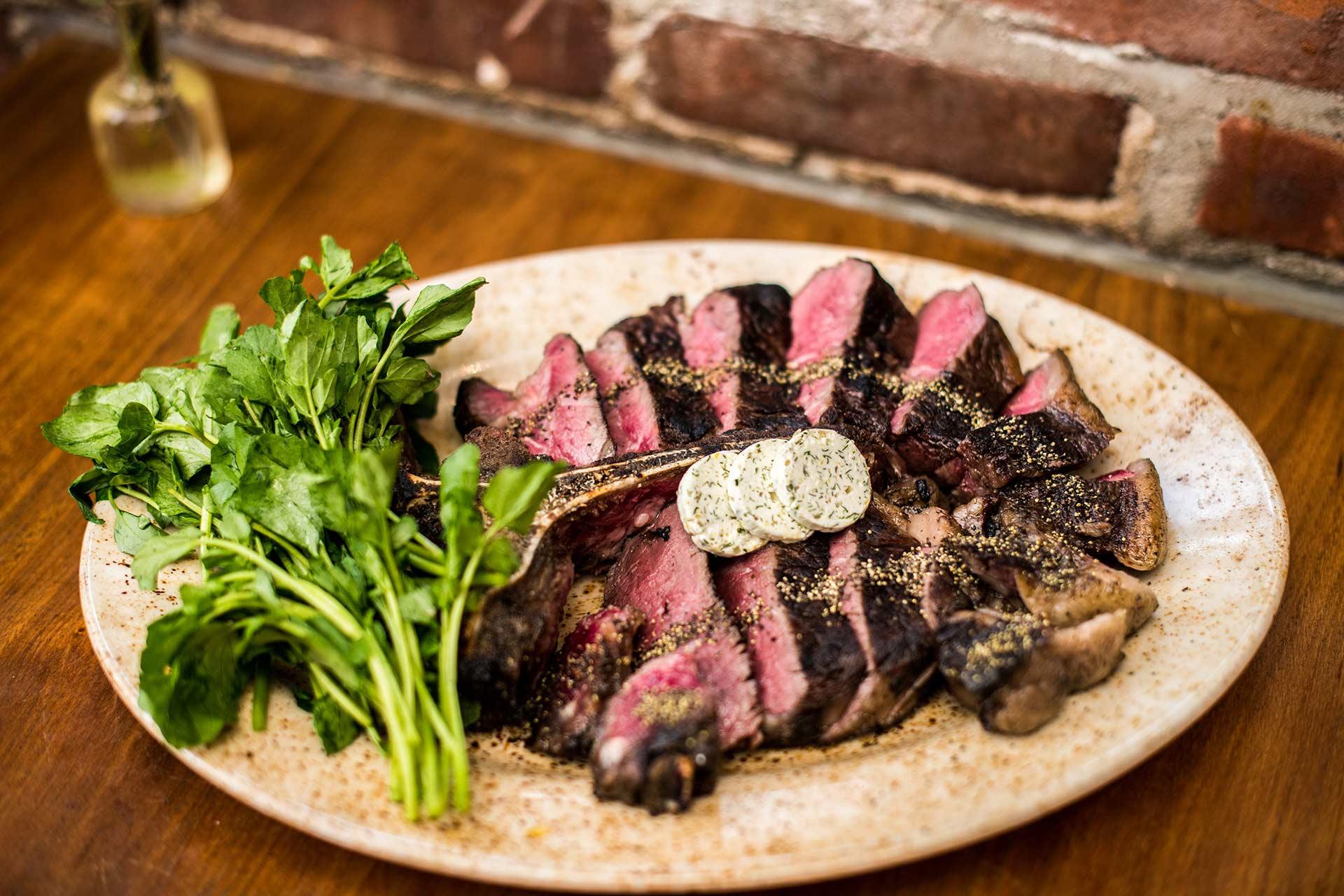 A plate of sliced, medium-rare steak with cracked pepper, topped with herb butter, served next to a bunch of fresh parsley on a rustic plate, set on a wooden table with a brick wall in the background.