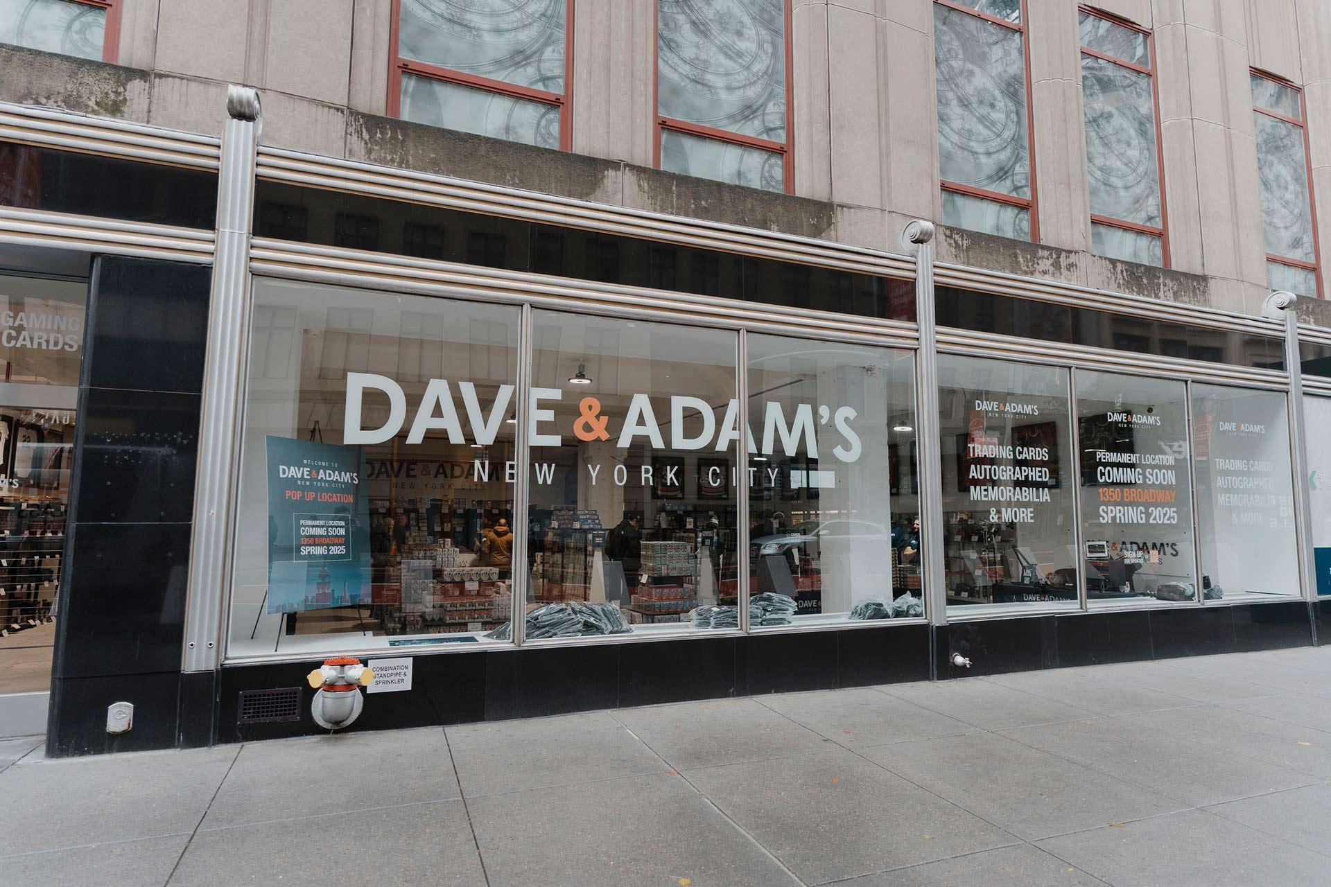 Storefront of Dave & Adam's in New York City, featuring large glass windows with signage promoting trading cards, memorabilia, and a movie section coming soon. Sparse street scene, with buildings reflected in the windows.