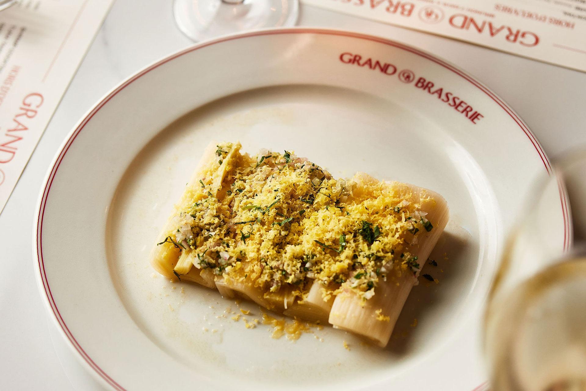A rectangular portion of pasta topped with grated cheese and herbs is served on a white plate labeled "GRAND BRASSERIE," with a wine glass and menu visible in the background.