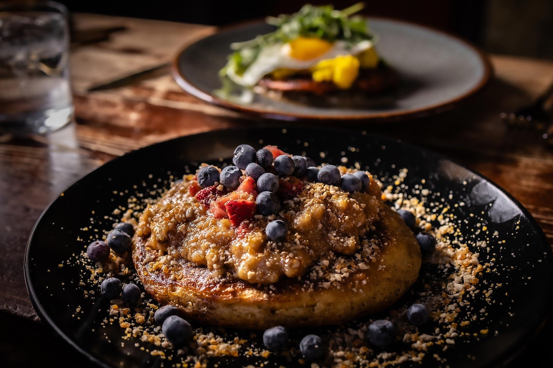 A close-up of a pancake topped with blueberry, nut crumble, and fruit compote on a dark plate, with a blurred dish of greens and an egg in the background on a rustic wooden table.